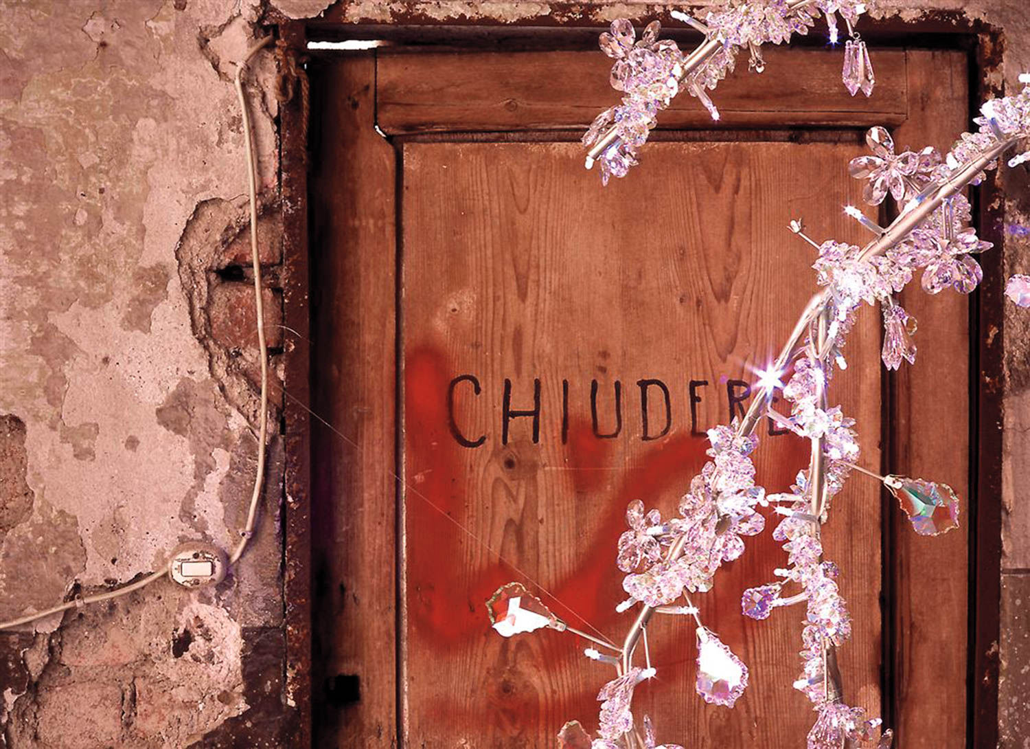 A weathered wooden door with “CHIUDERE” written on it, partially obscured by a decorative branch with clear crystals in the foreground.