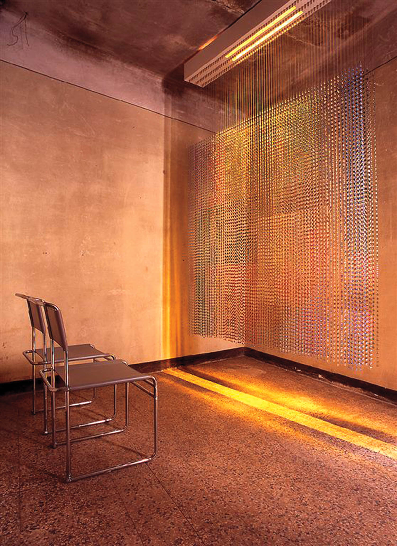 Three metal chairs face a wall in a bare room with warm light. Hanging beads or chains create a colorful, shimmering curtain effect near the corner.