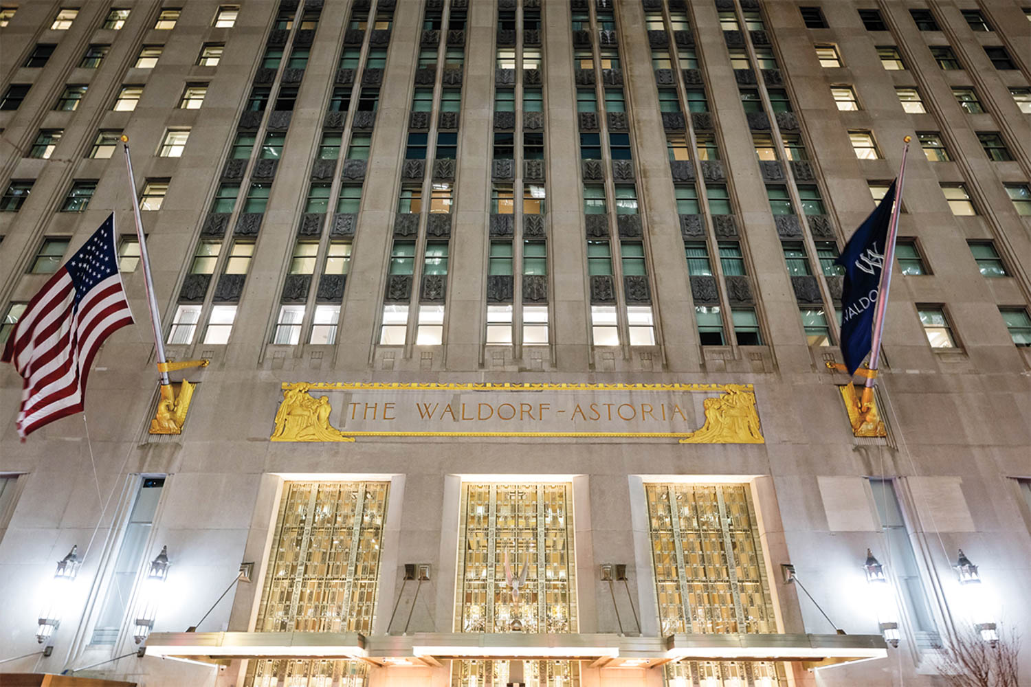 Facade of The Waldorf Astoria hotel with two flags, gold accents, and multiple lit windows at night.