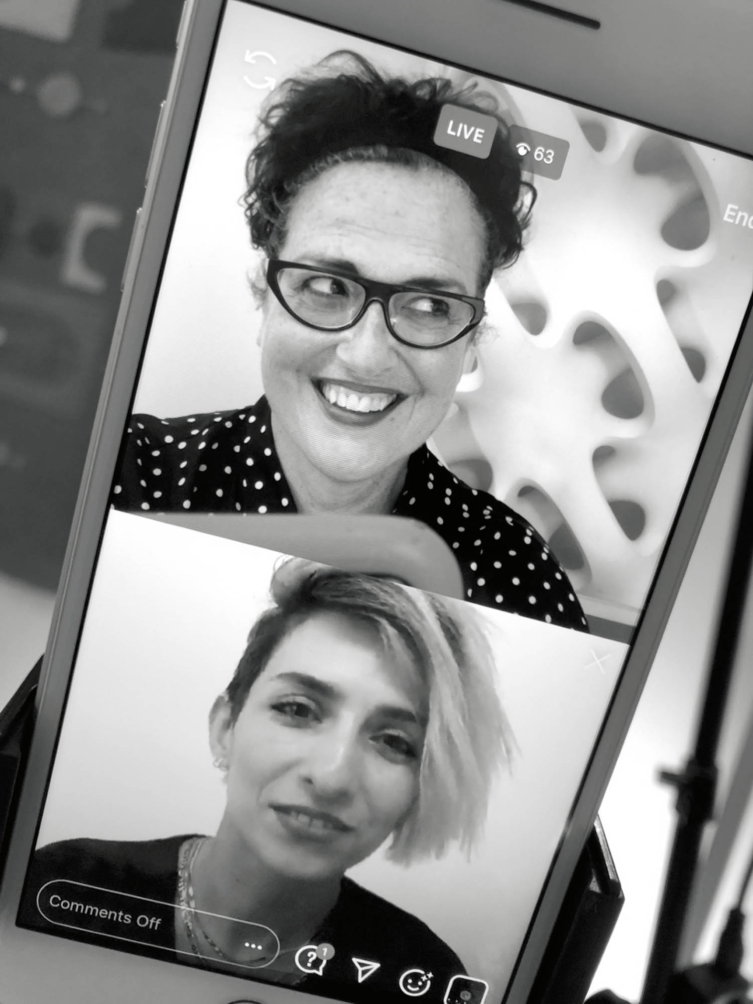 A phone screen shows two women in a split-screen video call; the top woman smiles and wears glasses, while the bottom woman has short, side-swept hair and looks at the camera.