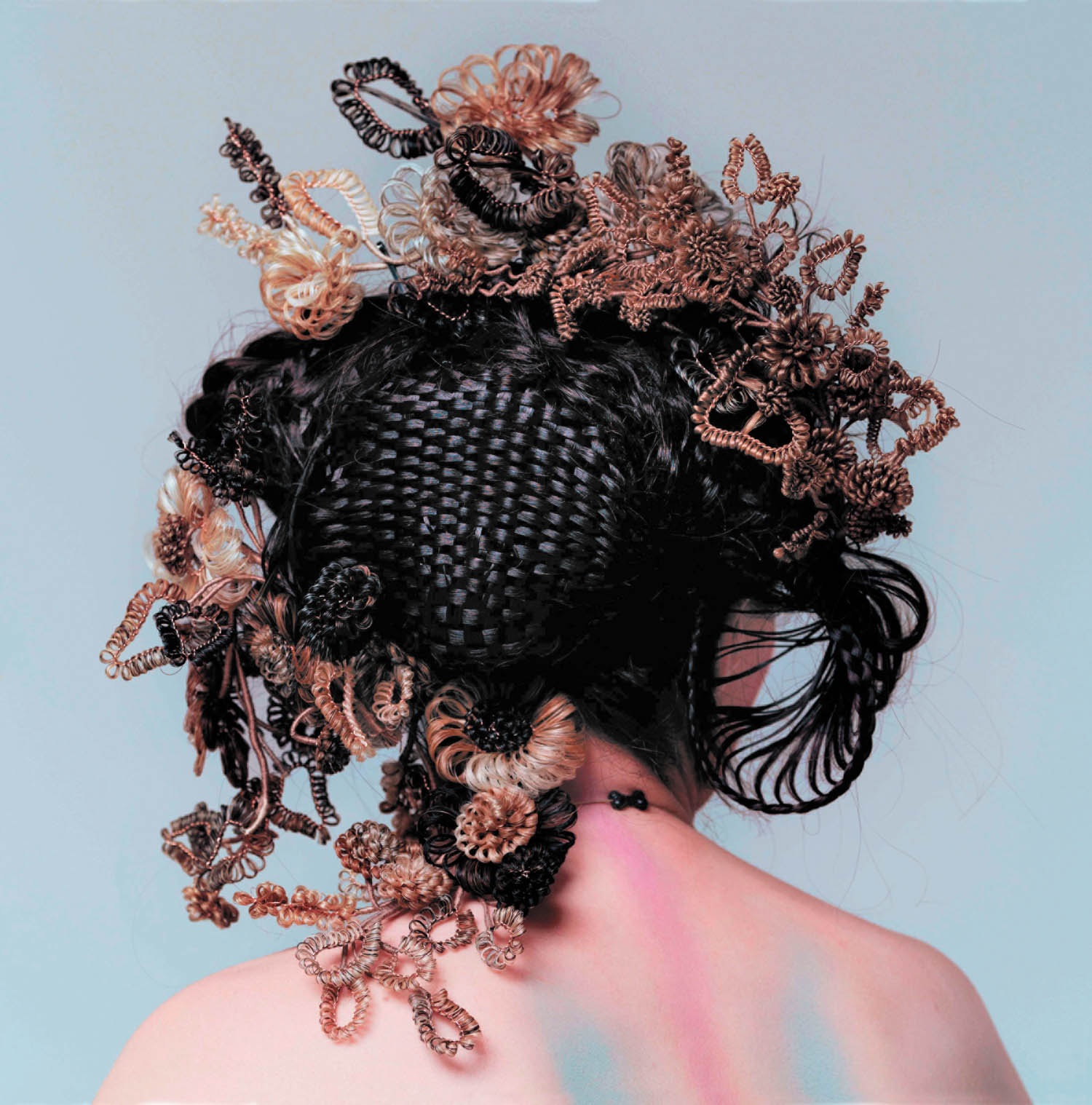 Person with their back to the camera, hair styled in intricate woven and braided patterns with added curly and looped hair decorations, on a light background.