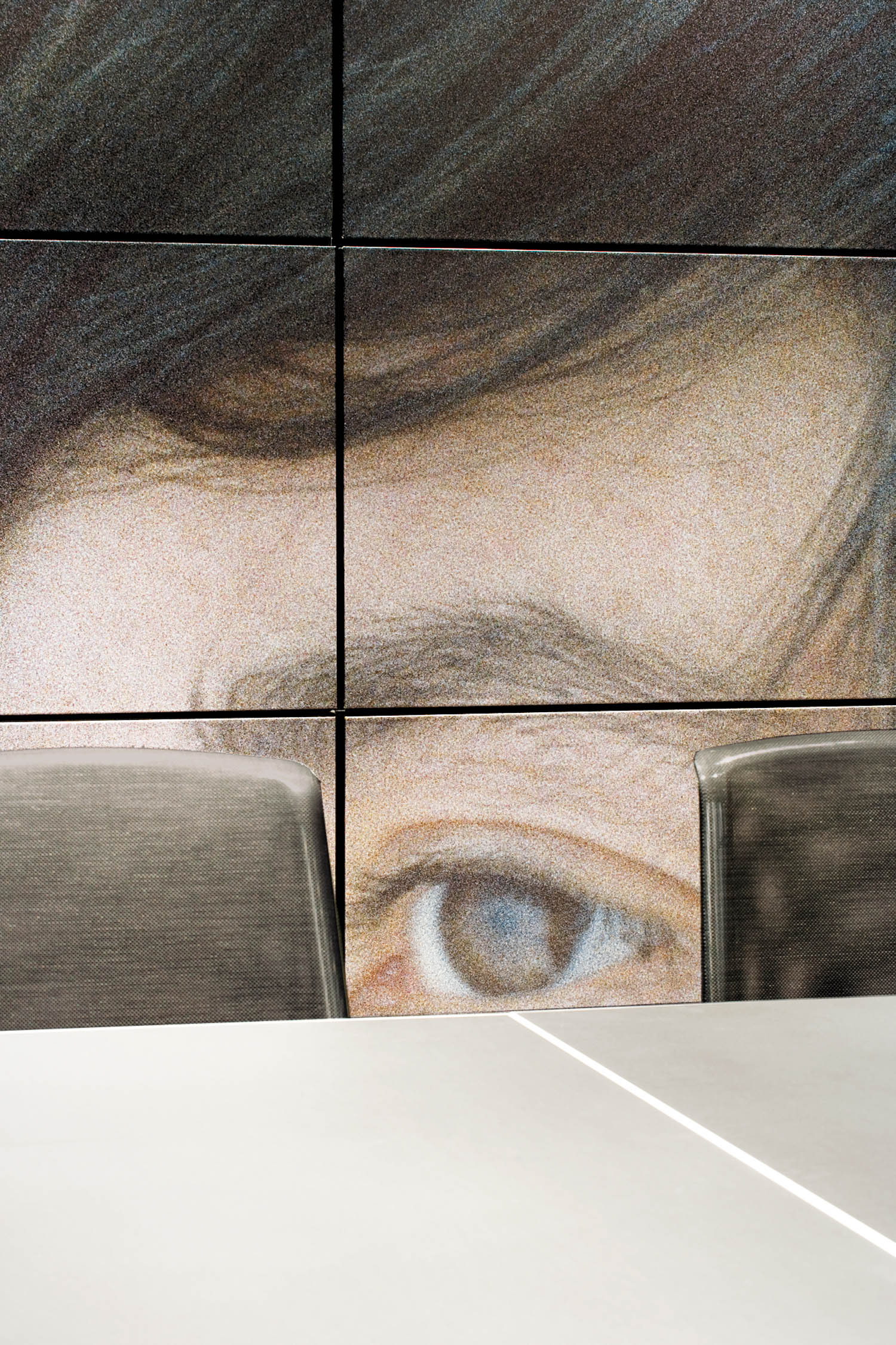 A large wall mural of a person’s eye and hair is partially visible behind two empty mesh chairs and a white table.