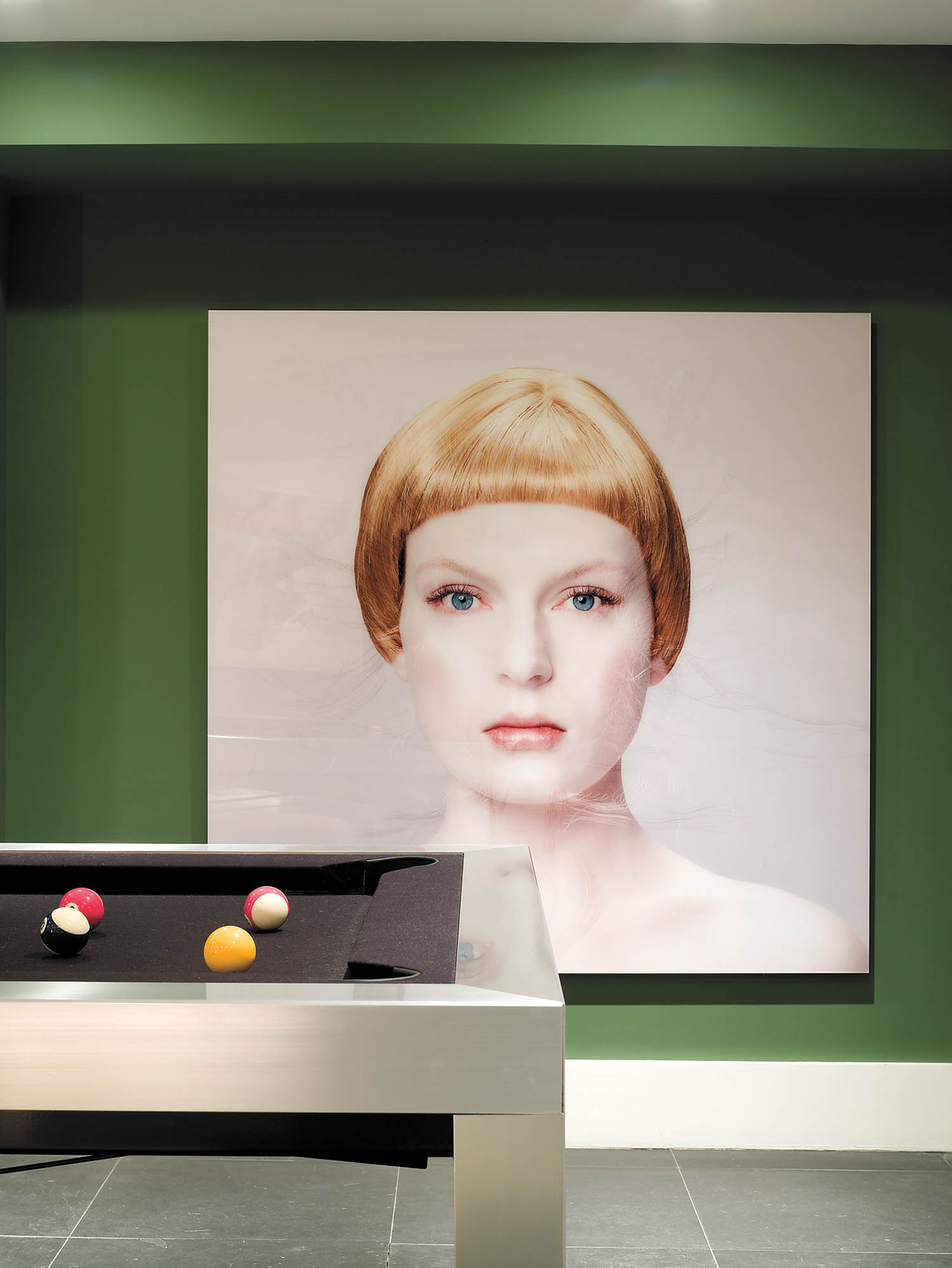 A wall features a large portrait of a woman with blond hair behind a modern pool table with four balls on its black felt surface.