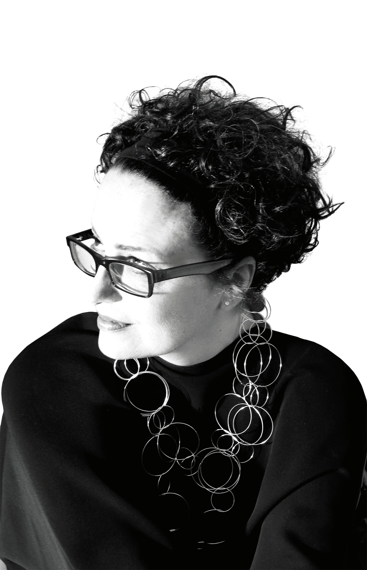 A person with short curly hair, glasses, and a large circular necklace looks to the side against a white background.