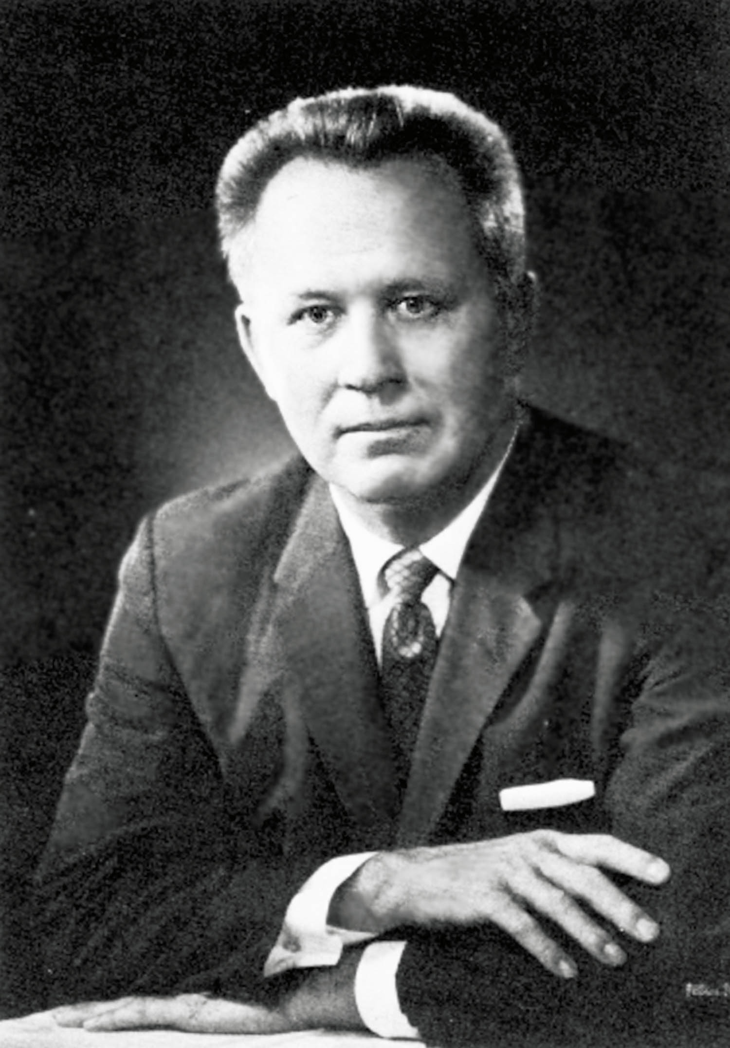 Black and white portrait of a middle-aged man in a suit and tie, seated with his arms crossed on a table, looking directly at the camera.