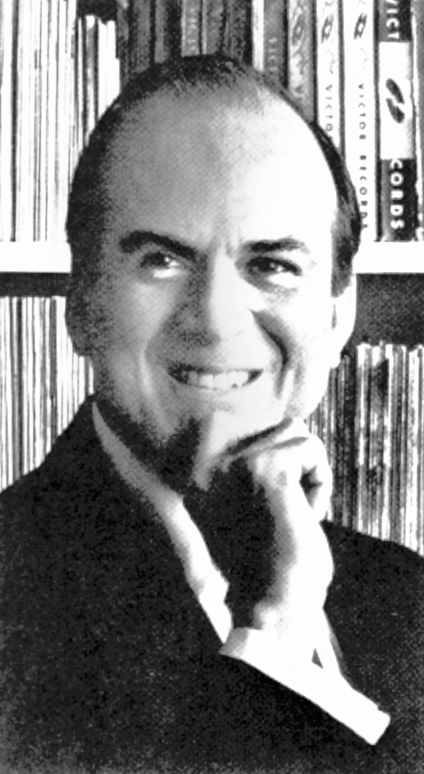 A man in a suit smiles at the camera with his hand resting on his chin, against a backdrop of bookshelves filled with books.