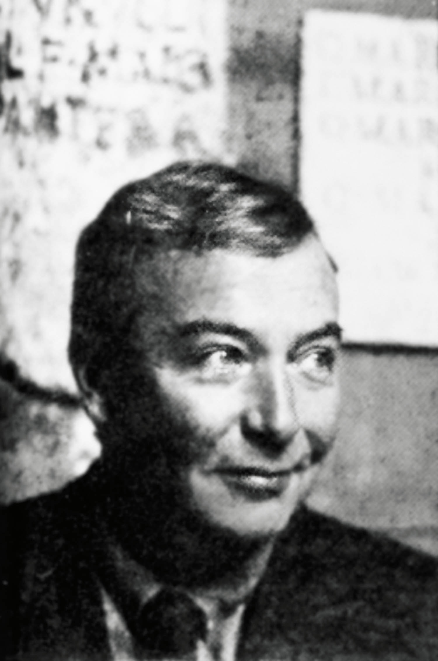 A man with short hair and a slight smile looks to the side. Blurry text is visible on the wall behind him. Black and white photo.