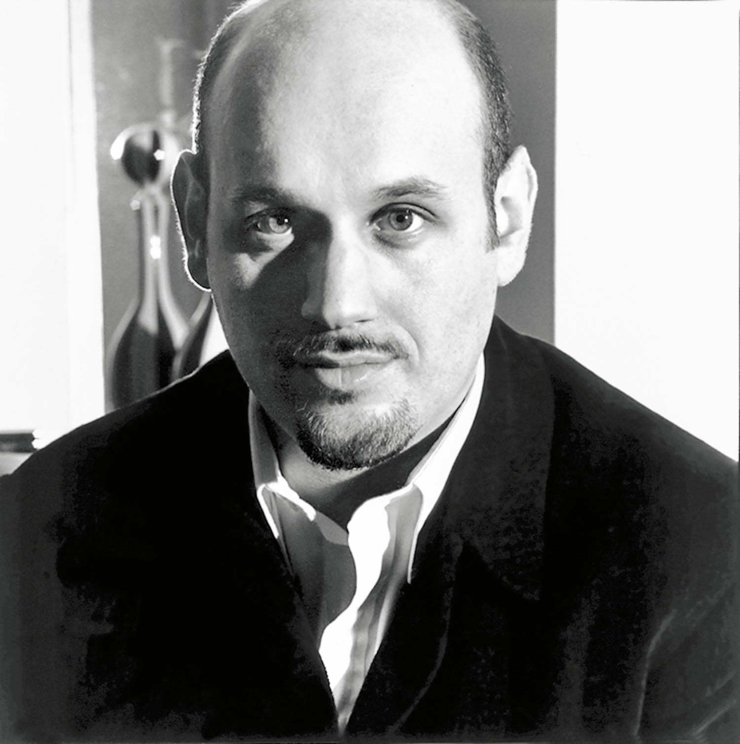 A bald man with a goatee, wearing a dark jacket over a white shirt, looks at the camera in a black and white portrait.