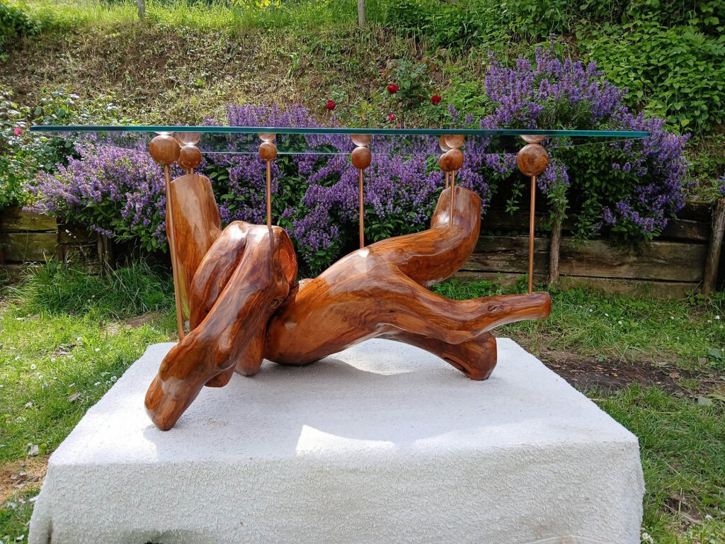 Abstract wooden sculpture displayed outdoors on a white platform, with purple flowers and greenery in the background. A metal rod with wooden spheres runs behind the sculpture.