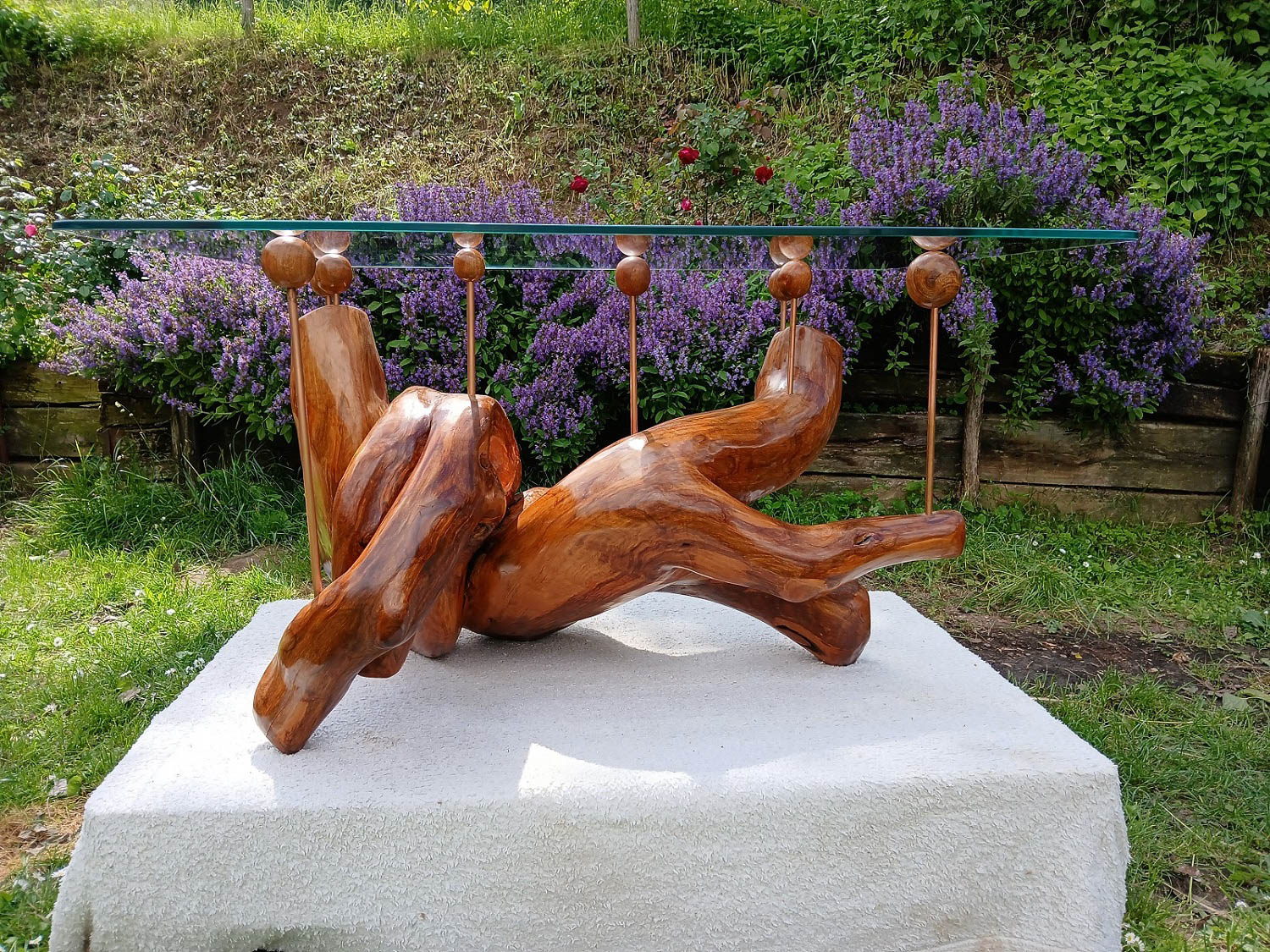 Abstract wooden sculpture displayed outdoors on a white platform, with purple flowers and greenery in the background. A metal rod with wooden spheres runs behind the sculpture.