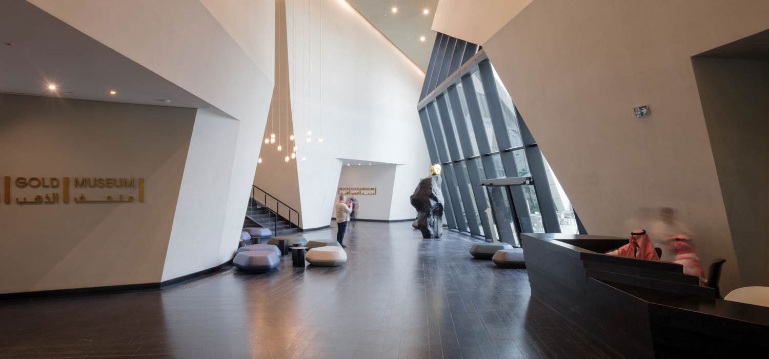 Modern museum lobby with angular white walls, large windows, dark floors reminiscent of black gold, reception desk to the right, and people walking or sitting throughout the space. Signs read “Gold Museum” and “Black Gold Museum” in English and Arabic.