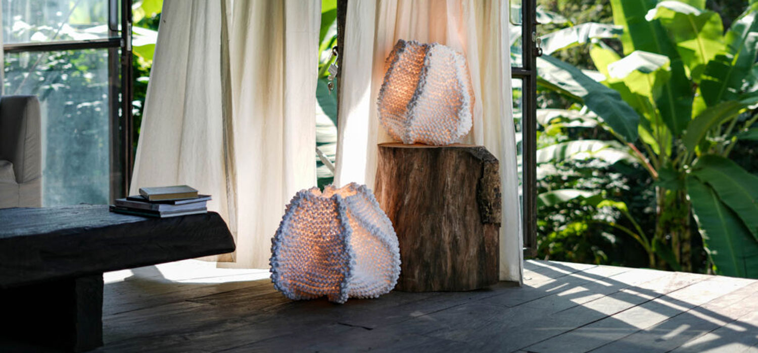 Two textured white lamps, one on a tree stump and one on the floor, are placed near sheer curtains with sunlight streaming in and lush green plants outside.