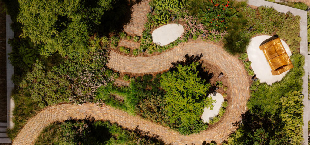 Curved brick path winds through a green garden with dense plants, two white circular areas, and a gold bench on a white platform.