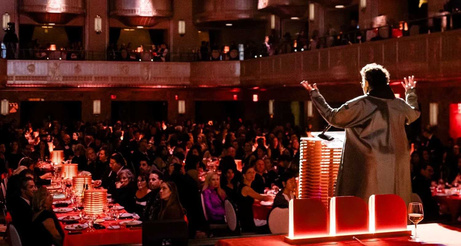 A speaker addresses a large seated audience in a banquet hall with red lighting and decorated tables.