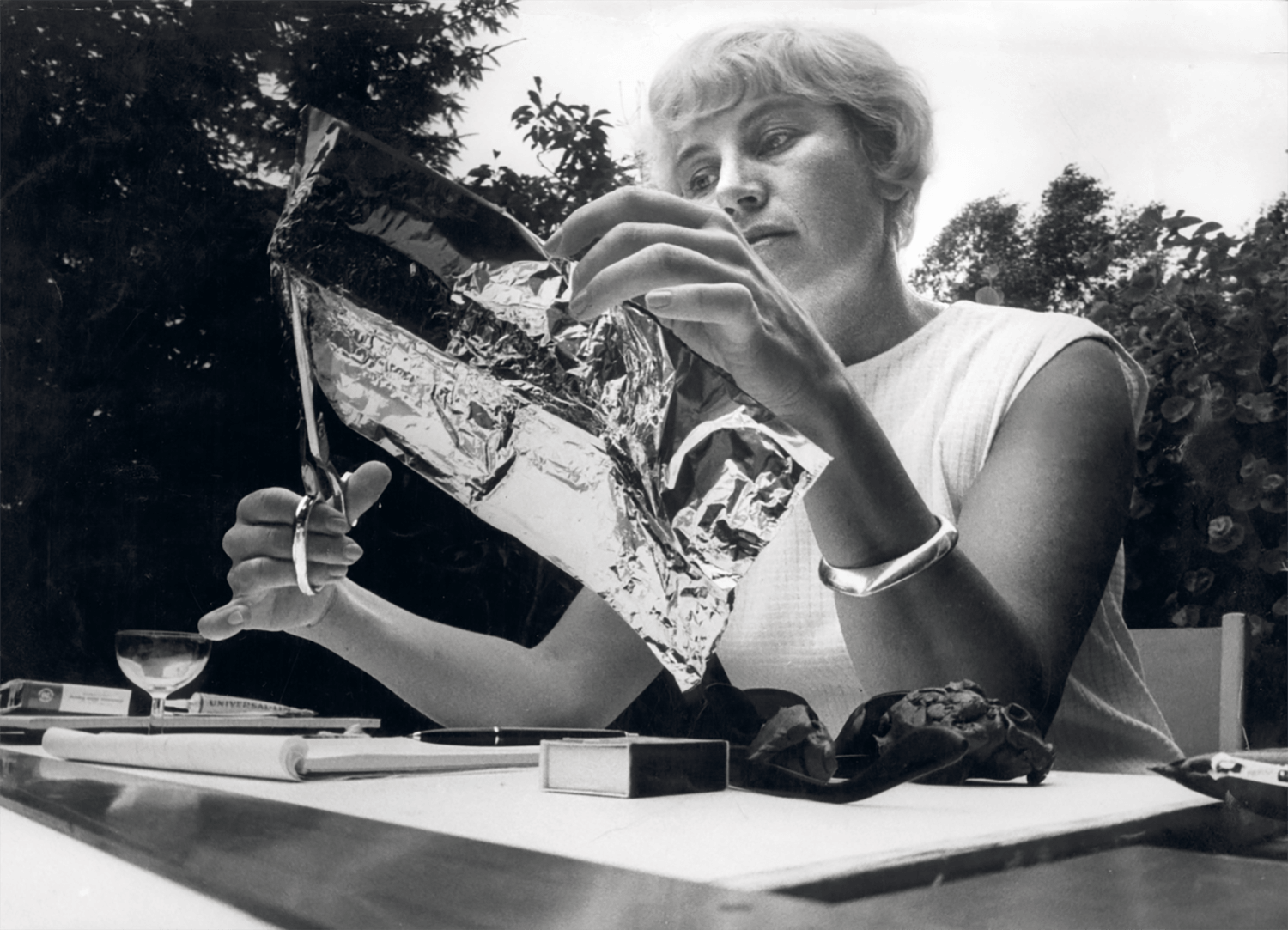 A person sits outdoors, cutting a sheet of reflective foil with scissors, surrounded by various objects on a table.