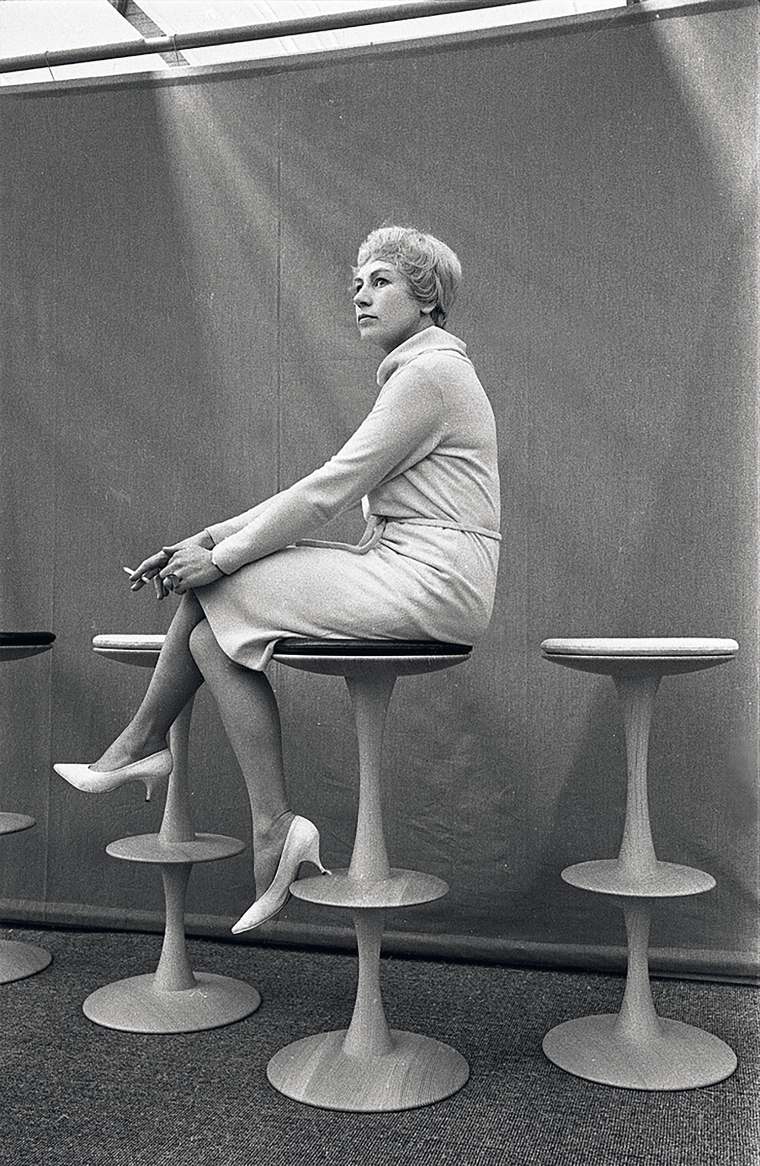 A woman in a light-colored outfit sits sideways on a tall modern stool, holding a cigarette, with similar stools beside her against a plain backdrop.