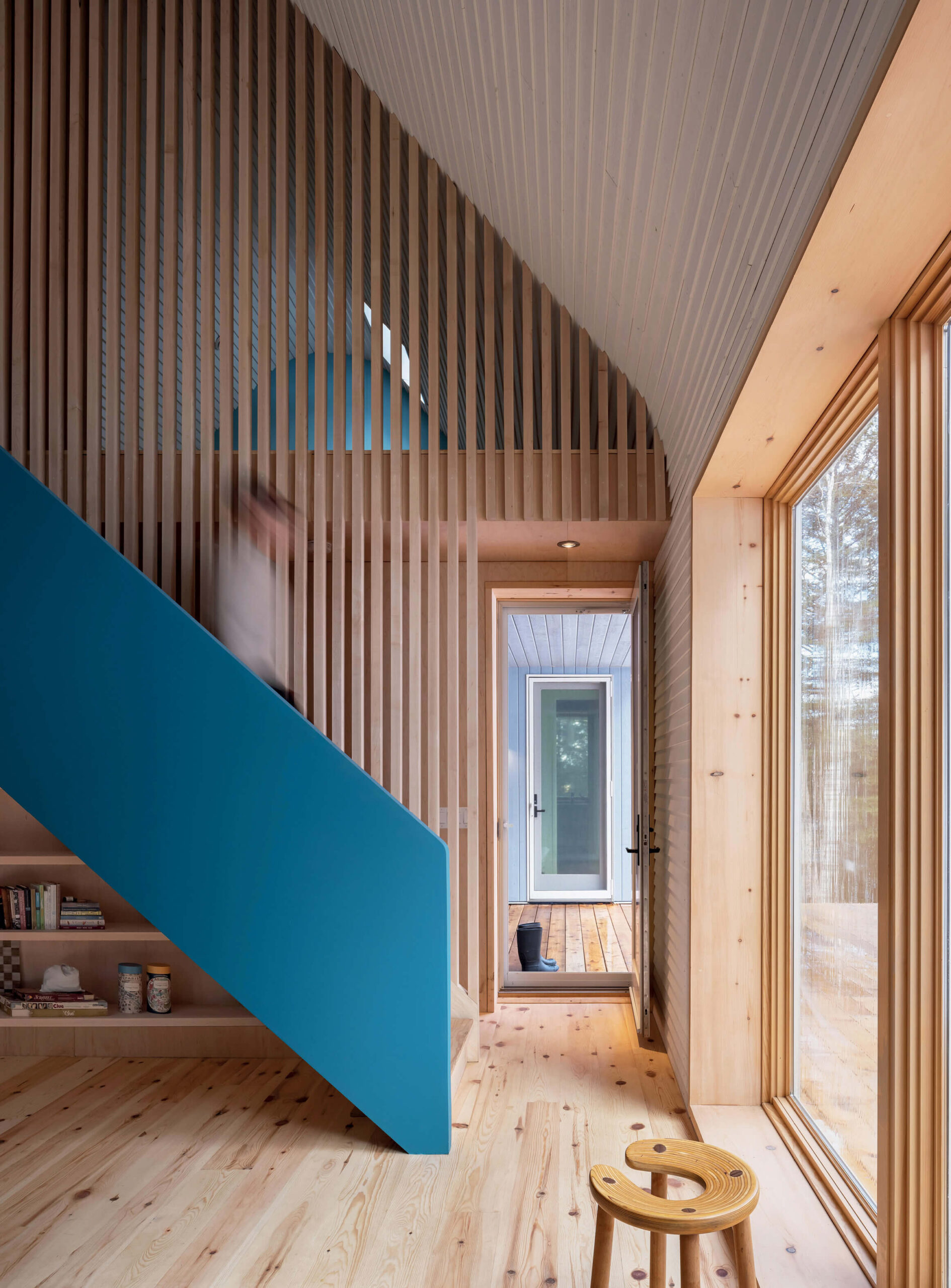 Modern wooden interior with large window, slatted walls, blue staircase, and a person ascending the stairs. A door leads outside; a stool and shelves are visible in the well-lit room.