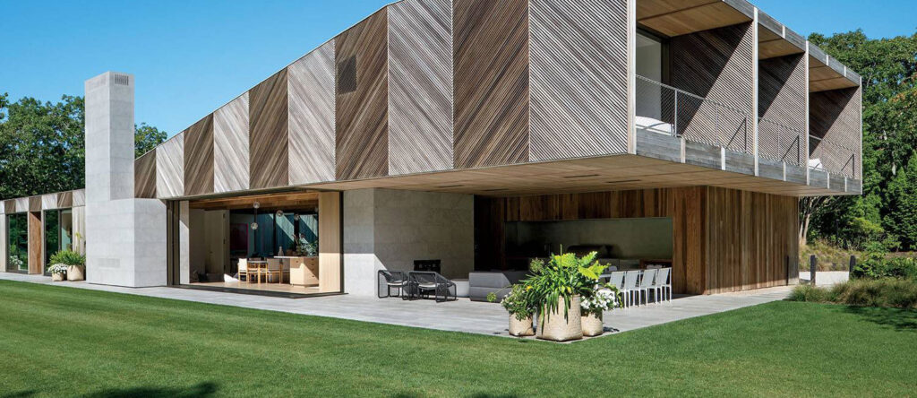 Modern two-story house with geometric wooden siding, large glass windows, outdoor seating areas, and a spacious grassy lawn.