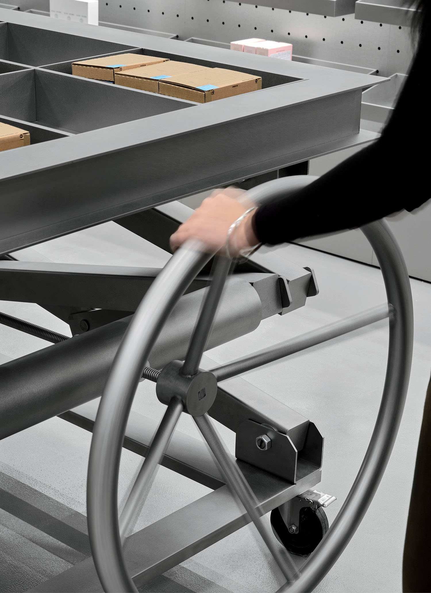 A person turns a large metal wheel attached to a gray industrial table with cardboard boxes on it in a modern, minimal workspace.