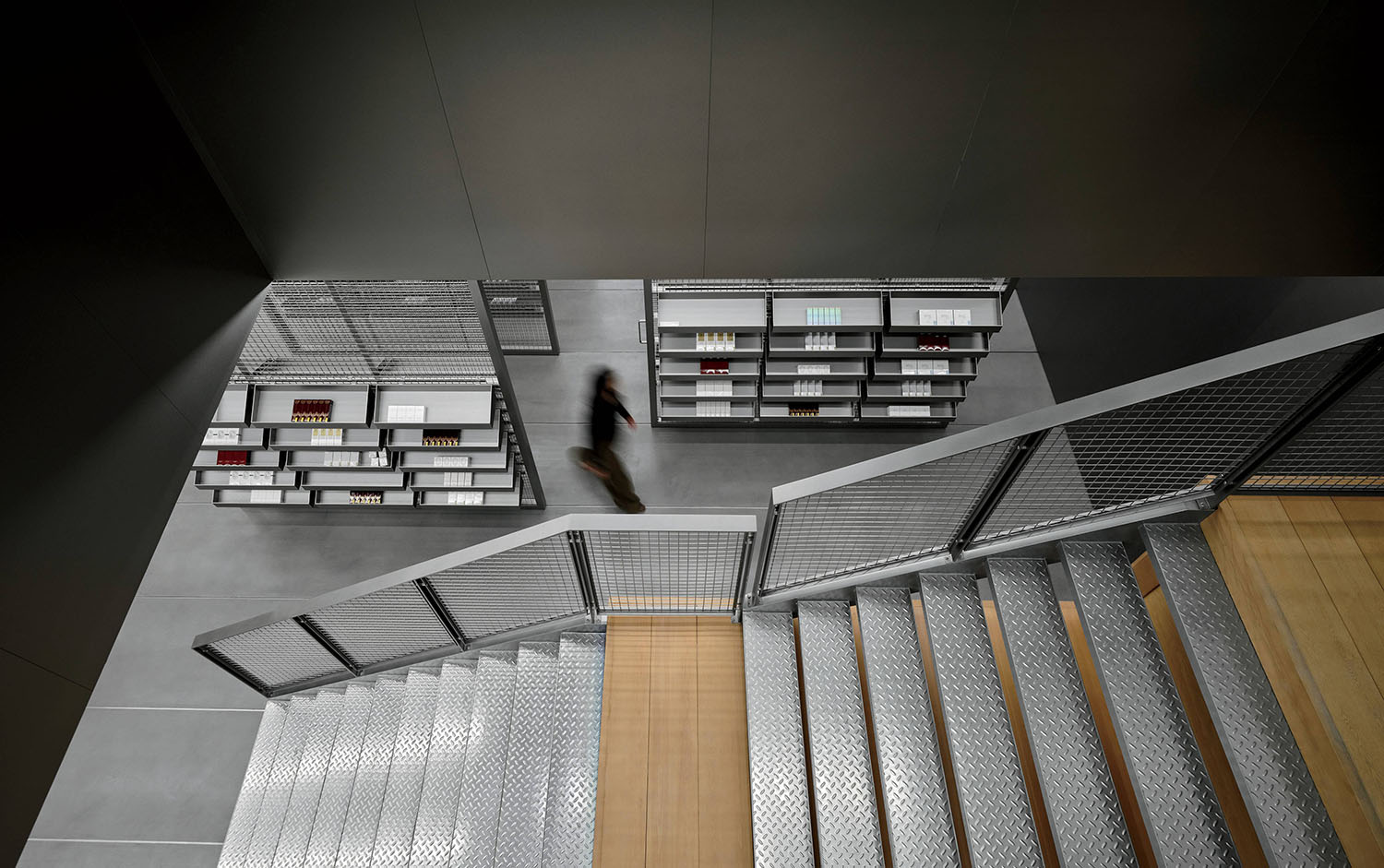 A person walks past shelves of books in a modern, industrial-style library viewed from the top of a metal staircase.