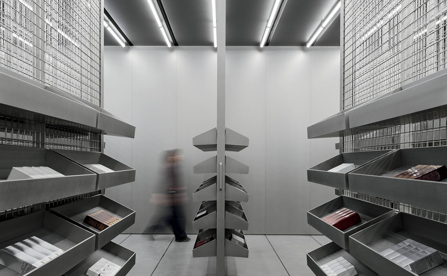 A person walks past metal shelving units filled with neatly stacked items in a modern, industrial-style room with bright ceiling lights.