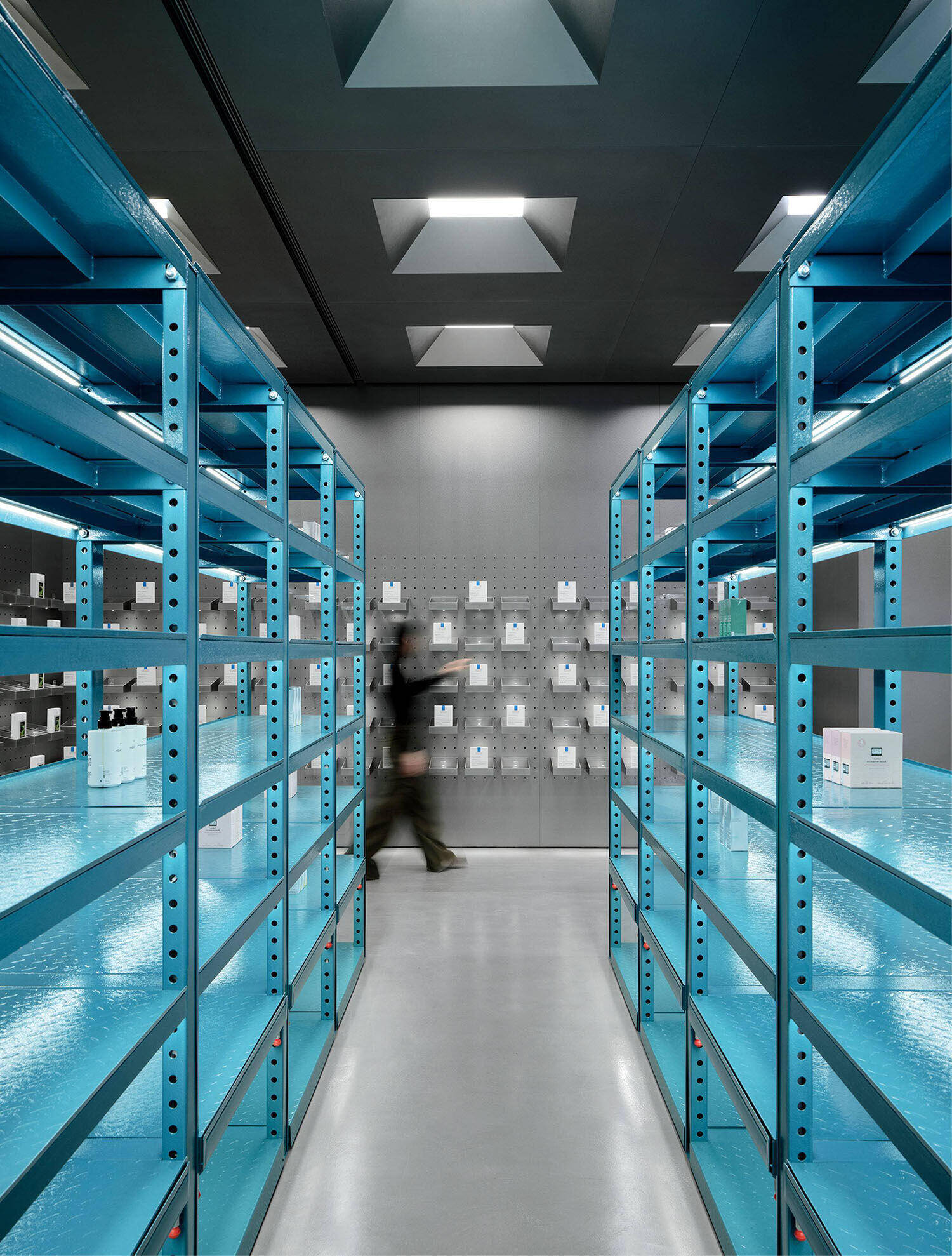 Person walking between blue metal shelves in a modern, industrial-style room with gray walls and boxed items displayed on pegs at the back.