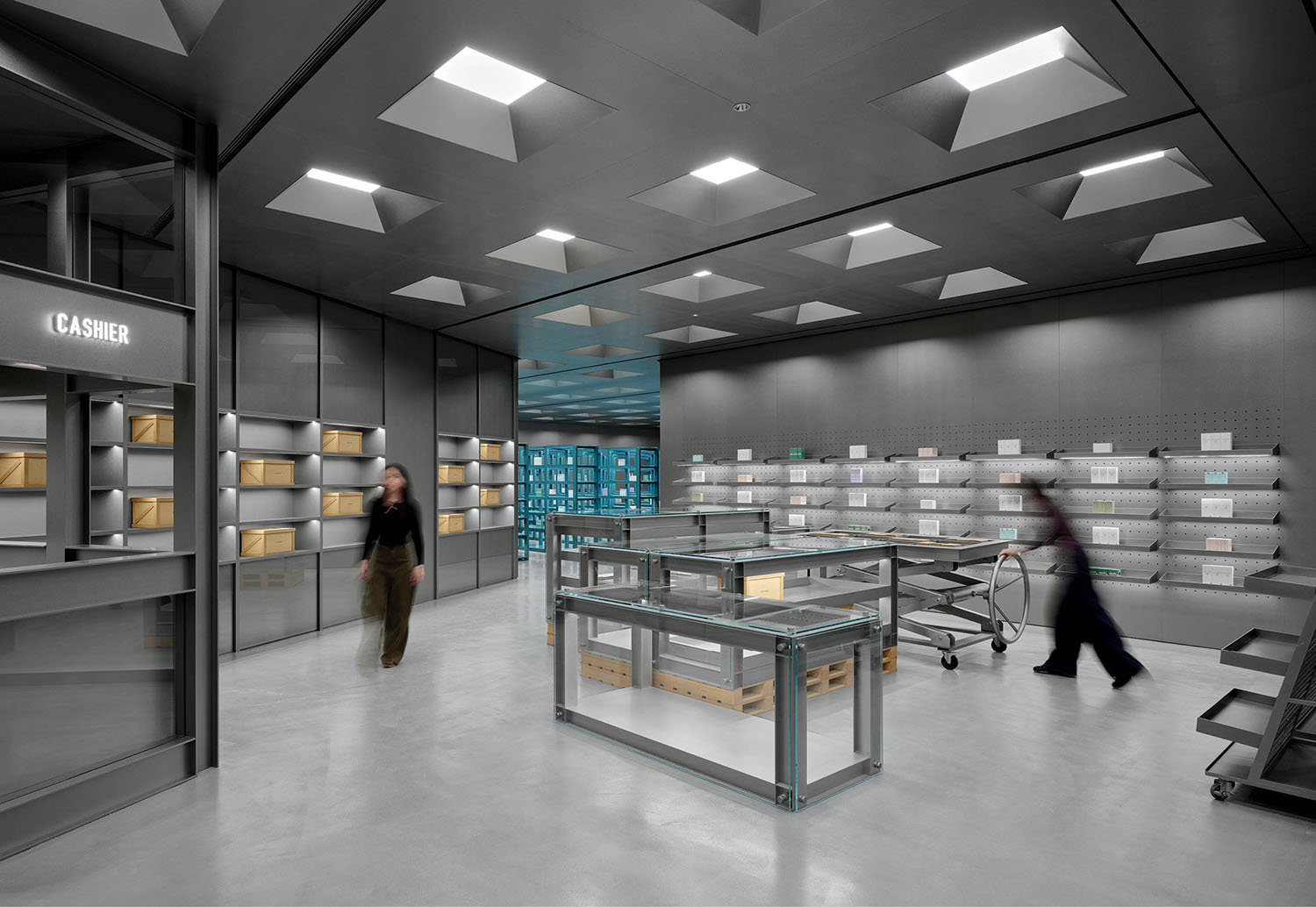 A modern, minimalist retail space with empty shelves, glass display tables, two people moving, and a cashier window on the left.