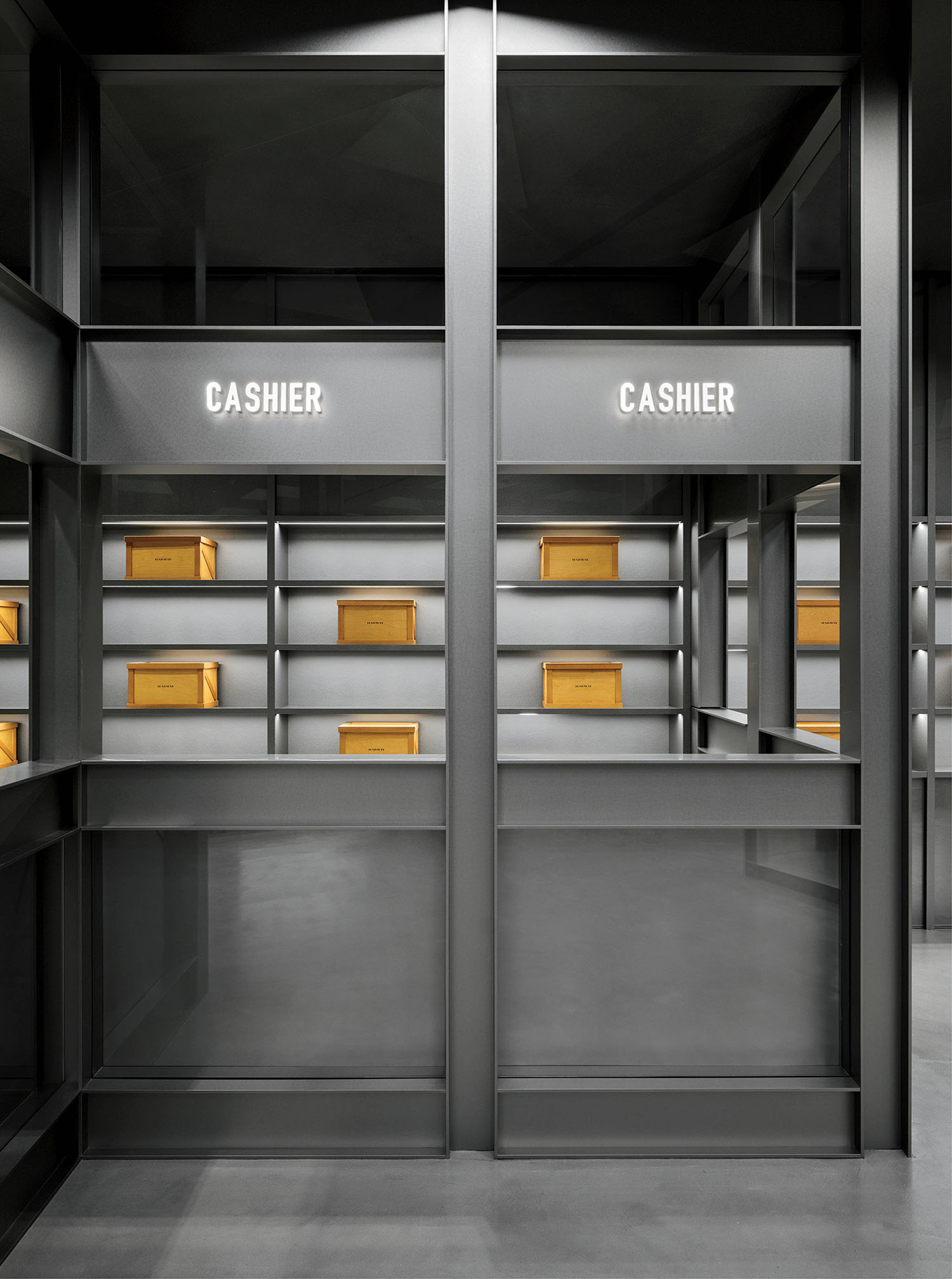 Minimalist cashier area with two labeled counters and shelves displaying several yellow boxes in a gray-toned interior.