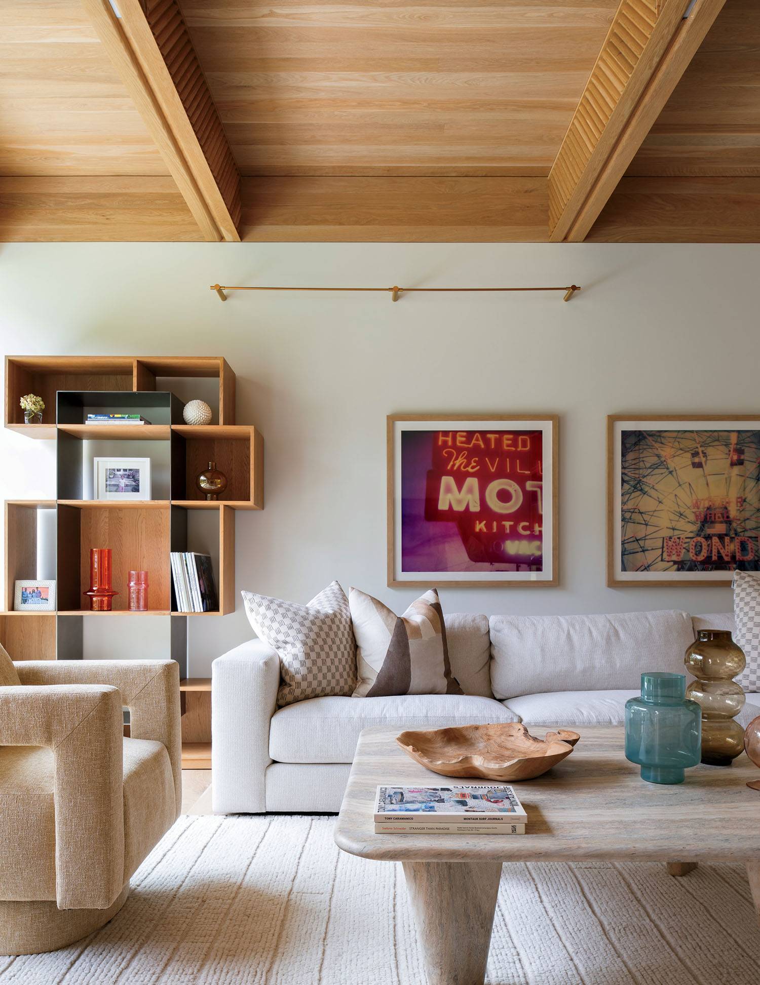 Modern living room with a beige sofa, patterned pillows, a wooden coffee table, shelving unit, framed wall art, and neutral decor under a wood beam ceiling.