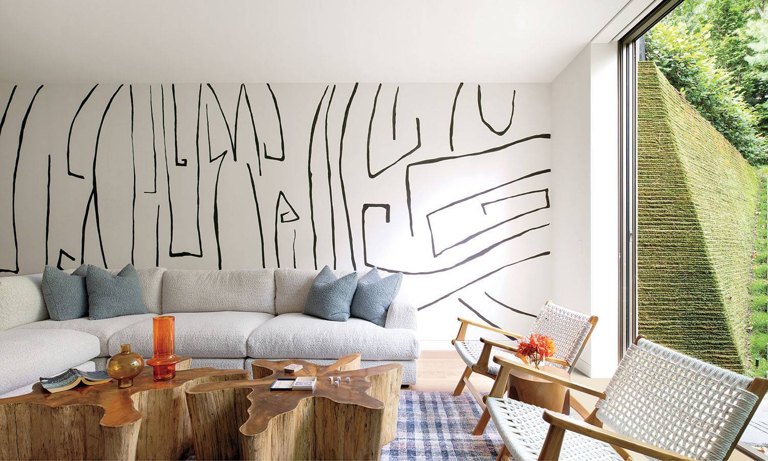 Modern living room with a large white sectional sofa, wood coffee tables, two chairs, abstract wall art, and floor-to-ceiling window showing a moss-covered exterior wall and greenery.