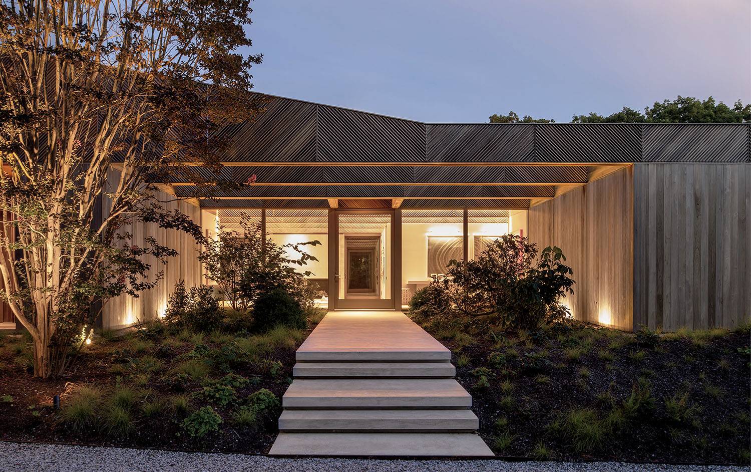 Modern house with wooden exterior, large front windows, and concrete steps leading to the glass front door, surrounded by landscaped plants and evening lighting.