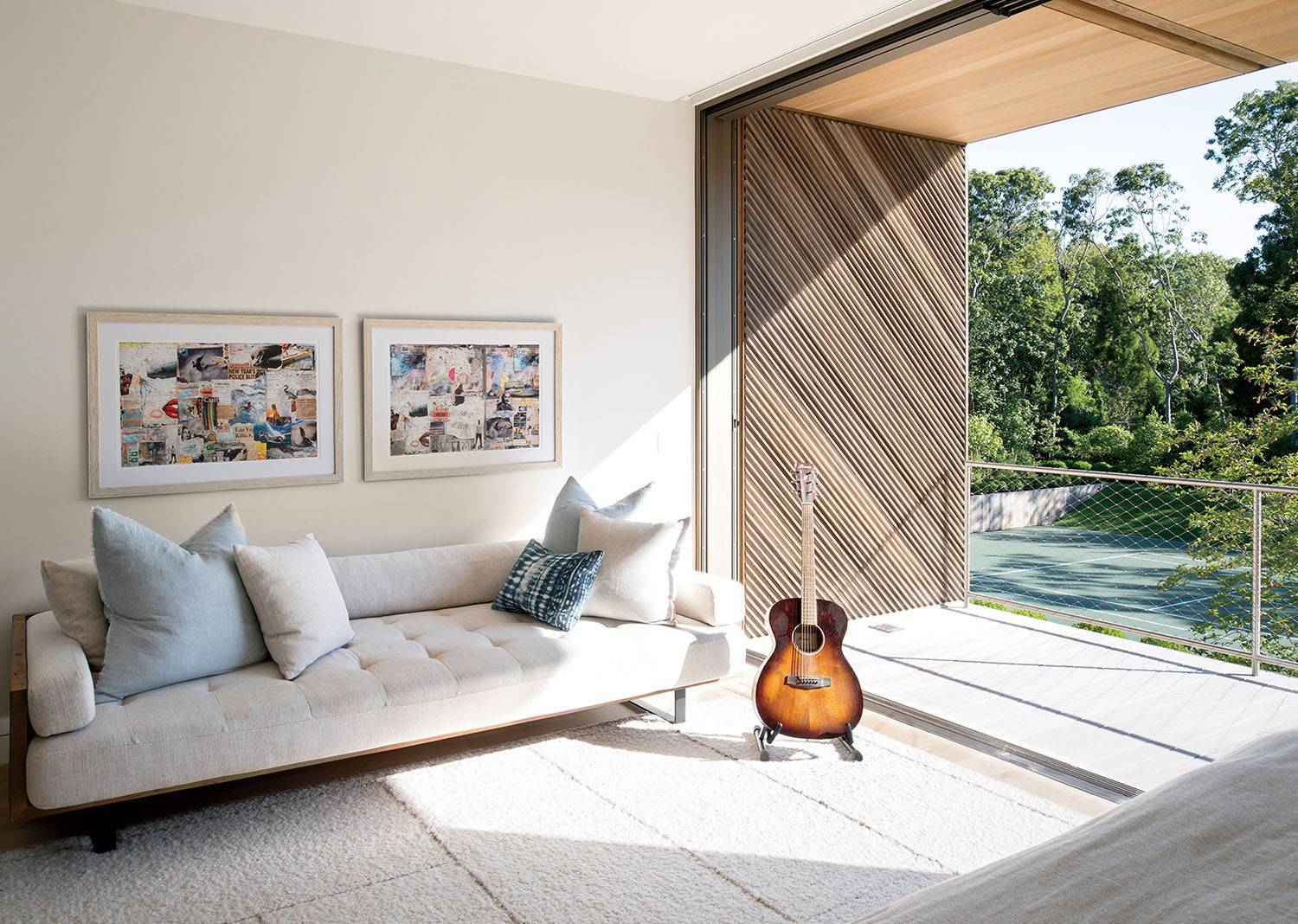 A sunlit modern living room with a white sofa, two framed artworks, a guitar on a stand, and a large sliding door open to a balcony with a view of trees.