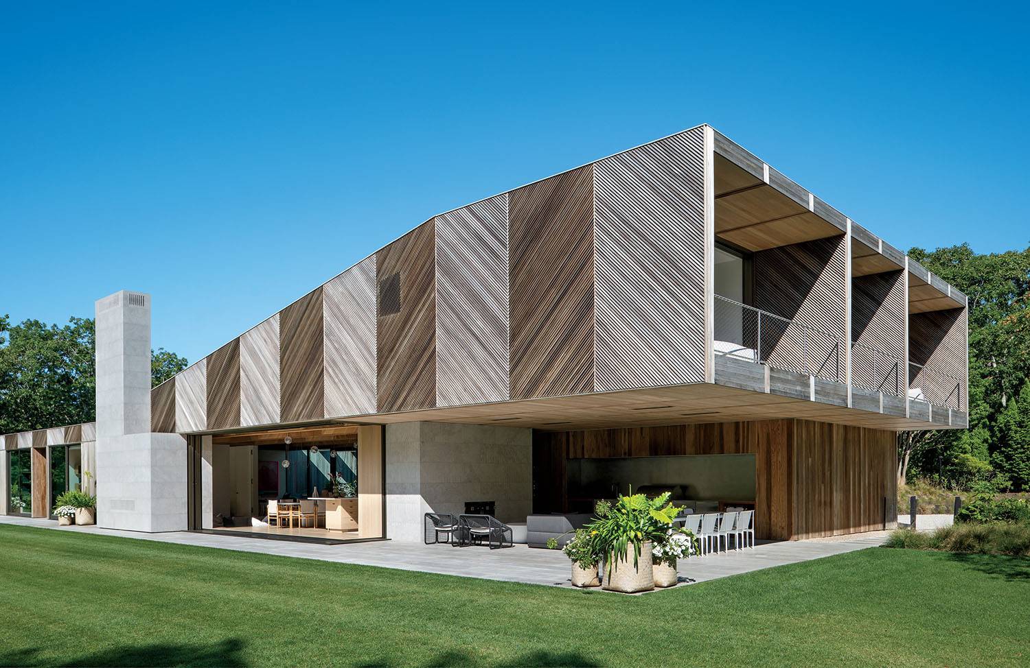 Modern two-story house with geometric design, wood and concrete exterior, large windows, covered patio, outdoor dining area, and a green lawn under a clear blue sky.