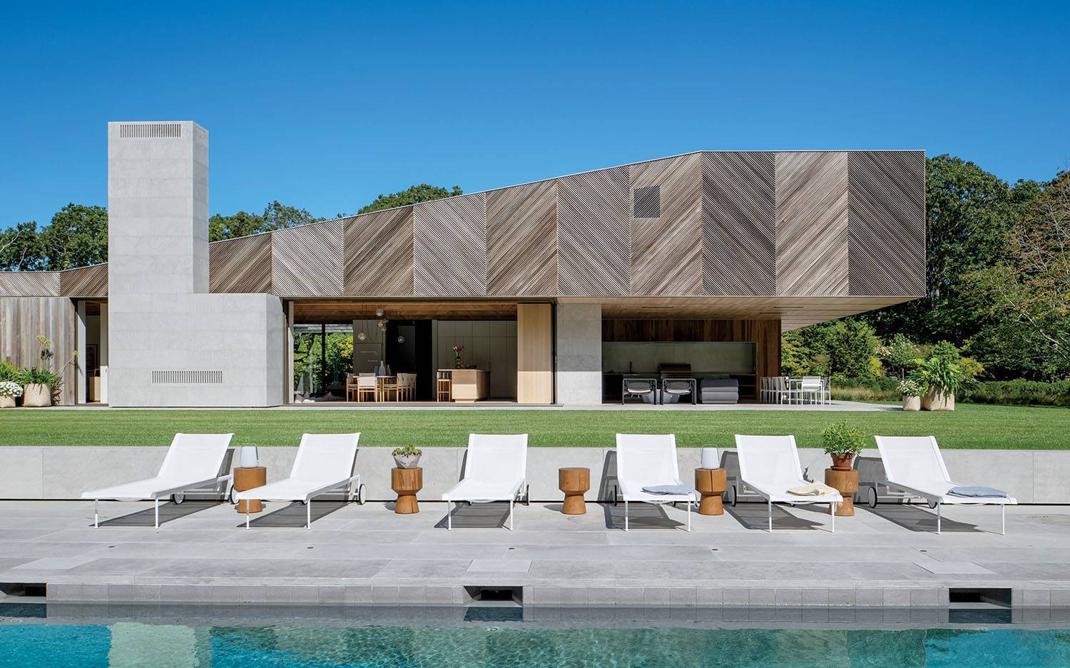 Modern house with geometric wood and concrete exterior, open patio, lawn, and row of white lounge chairs by a pool under a clear blue sky.