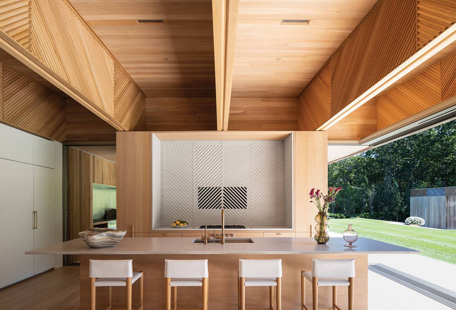 Modern kitchen with wooden cabinetry, large island with four stools, built-in stove, and sliding glass doors opening to a green outdoor area.