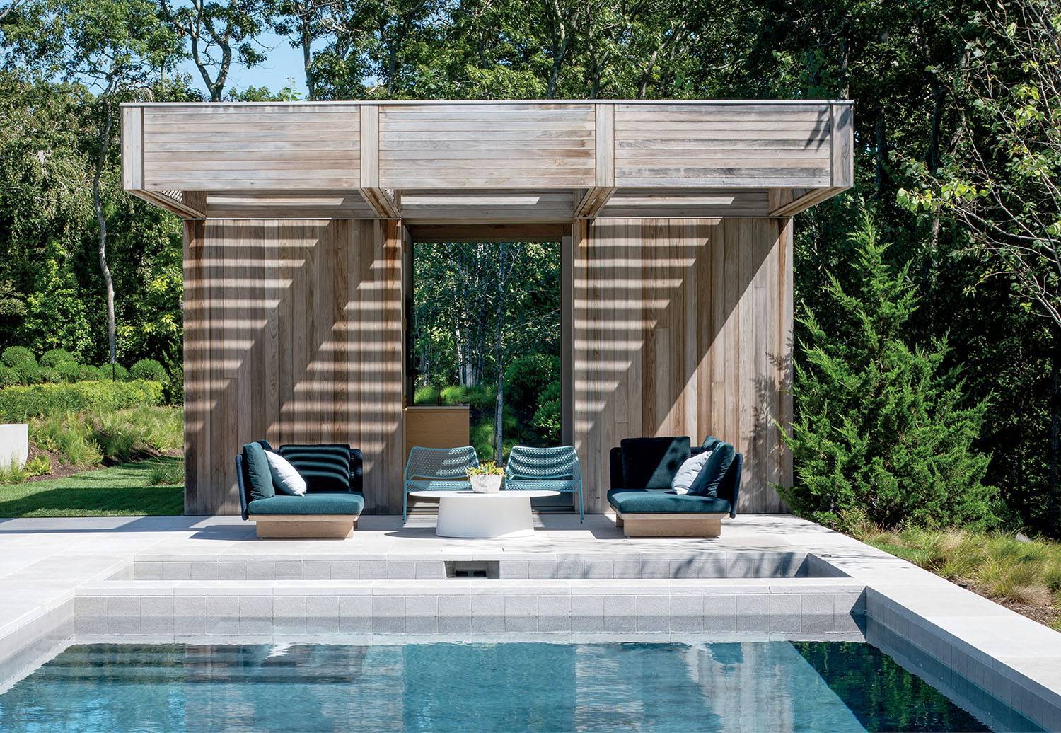 Modern wooden poolside cabana with lounge chairs and a table, set beside a rectangular swimming pool, surrounded by green trees and landscaping.