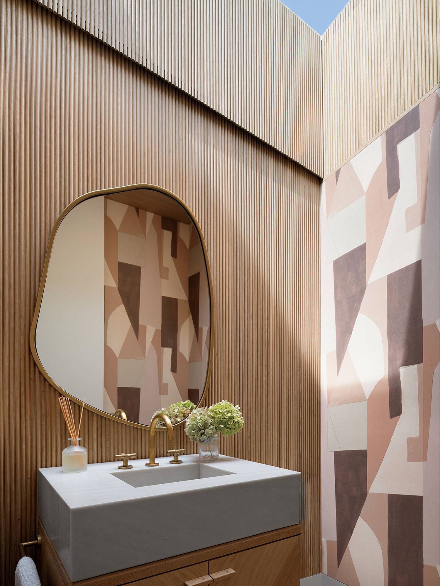 Modern bathroom with wood paneling, abstract geometric wallpaper, an irregular oval mirror, a rectangular sink, gold faucet, and a small vase with green flowers on the countertop.