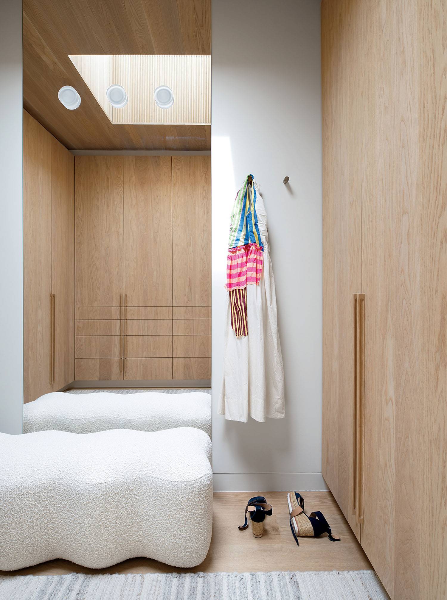 A minimalist walk-in closet with wooden cabinets, a skylight, a white textured bench, a dress and scarf on a hook, and two pairs of shoes on the floor.