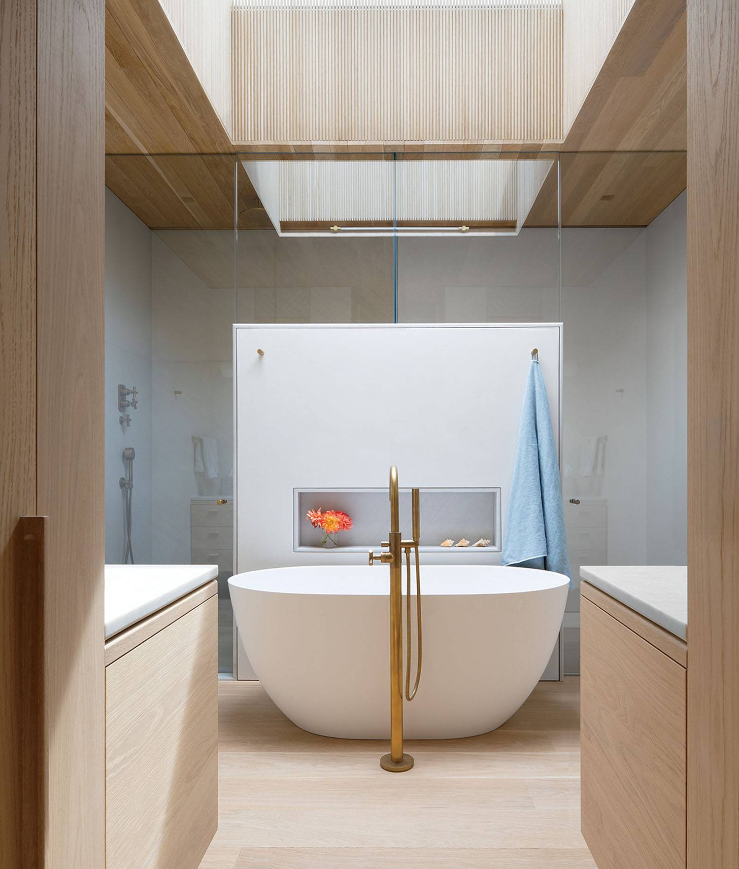 Minimalist bathroom with a freestanding white bathtub, brass fixtures, a light blue towel, built-in shelf, and natural wood accents under a skylight.