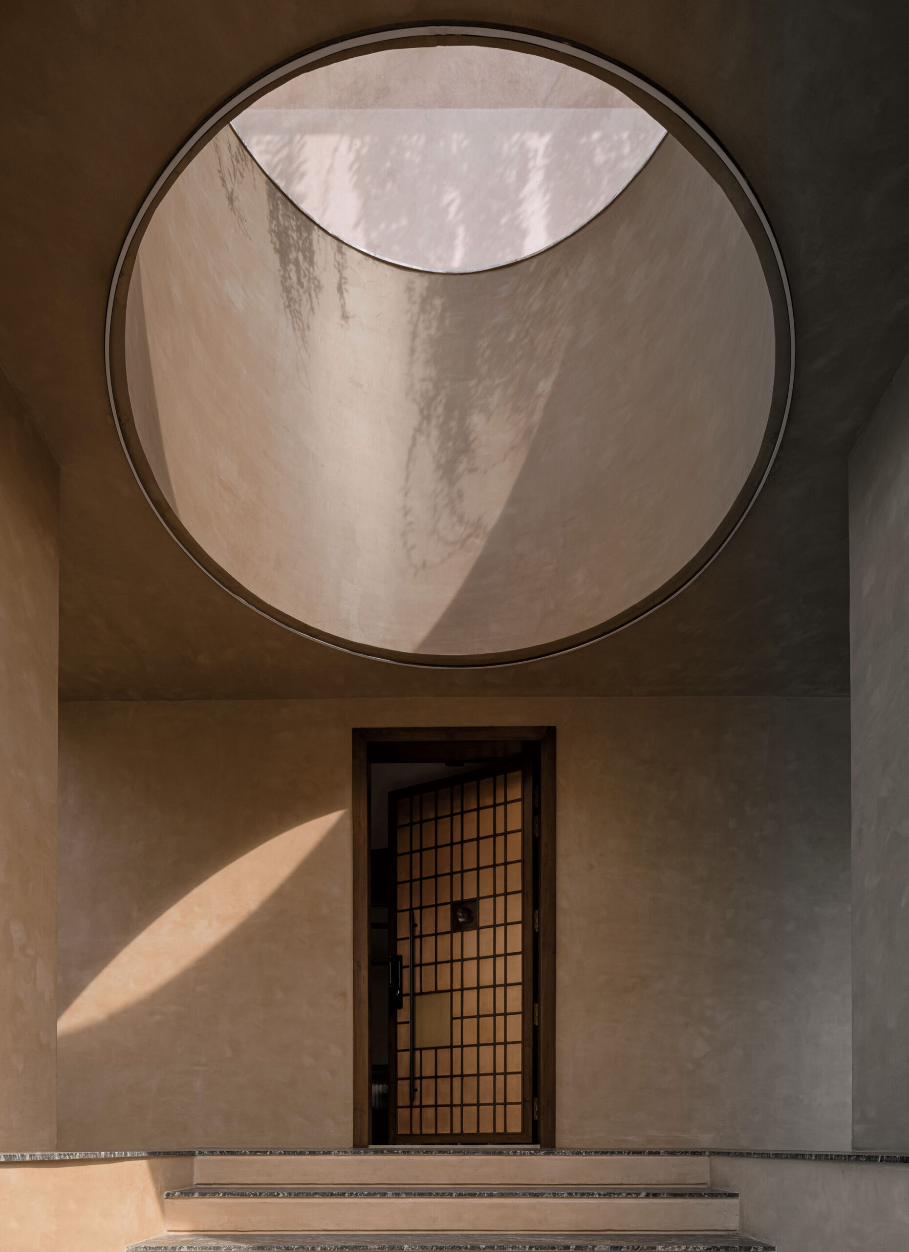 A geometric entryway with a circular skylight above a door featuring a grid pattern, casting natural light and shadows on beige walls and steps.