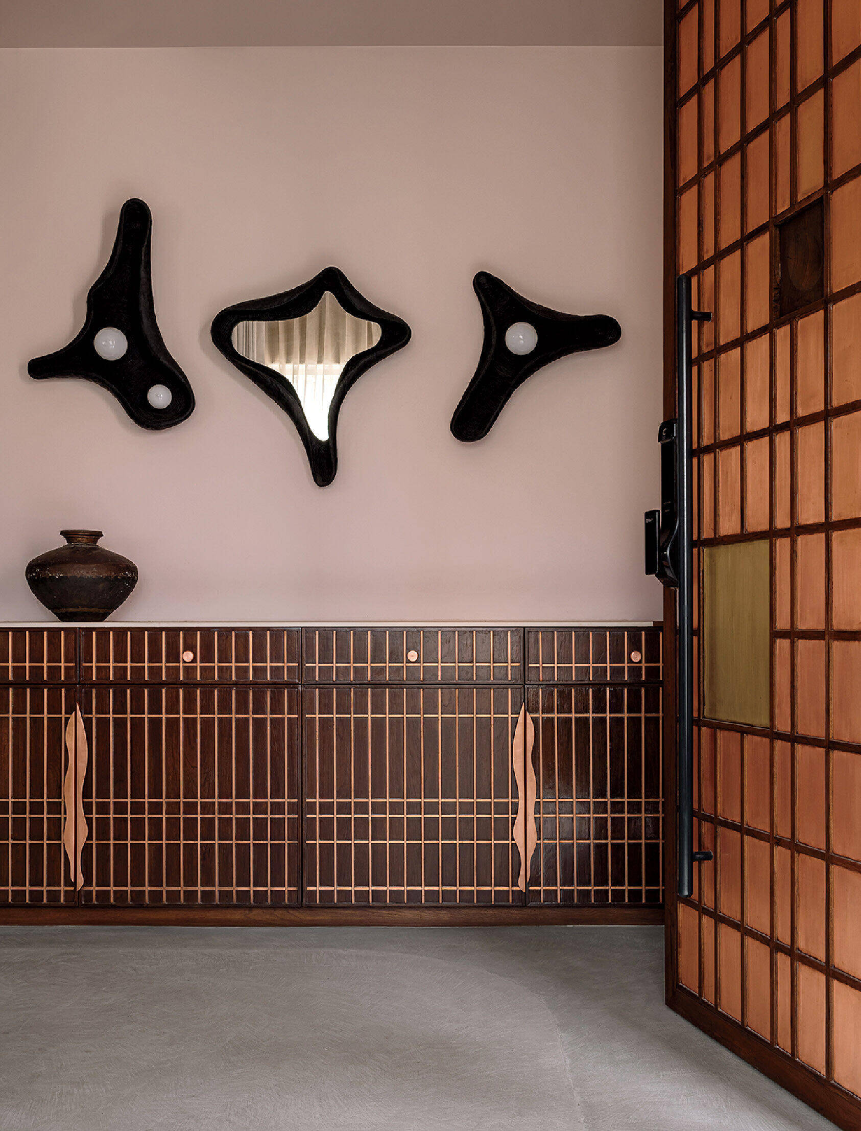 A minimalist room with geometric wooden cabinets, three abstract black wall mirrors, a ceramic vase, and a wooden lattice sliding door.
