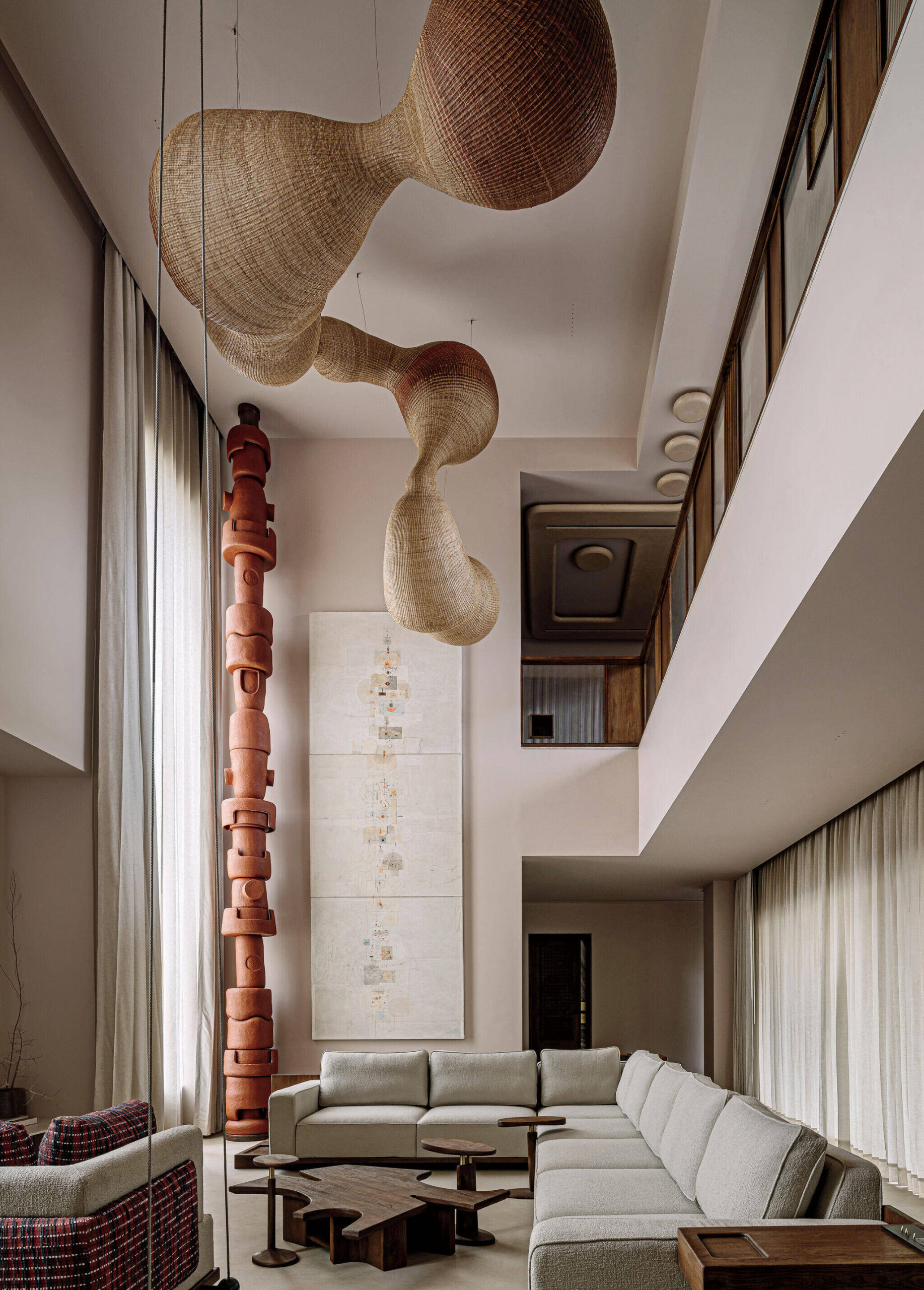 Modern living room with a large beige sectional sofa, sculptural wooden coffee tables, pendant woven lights, tall red art installation, neutral walls, and floor-to-ceiling curtains.