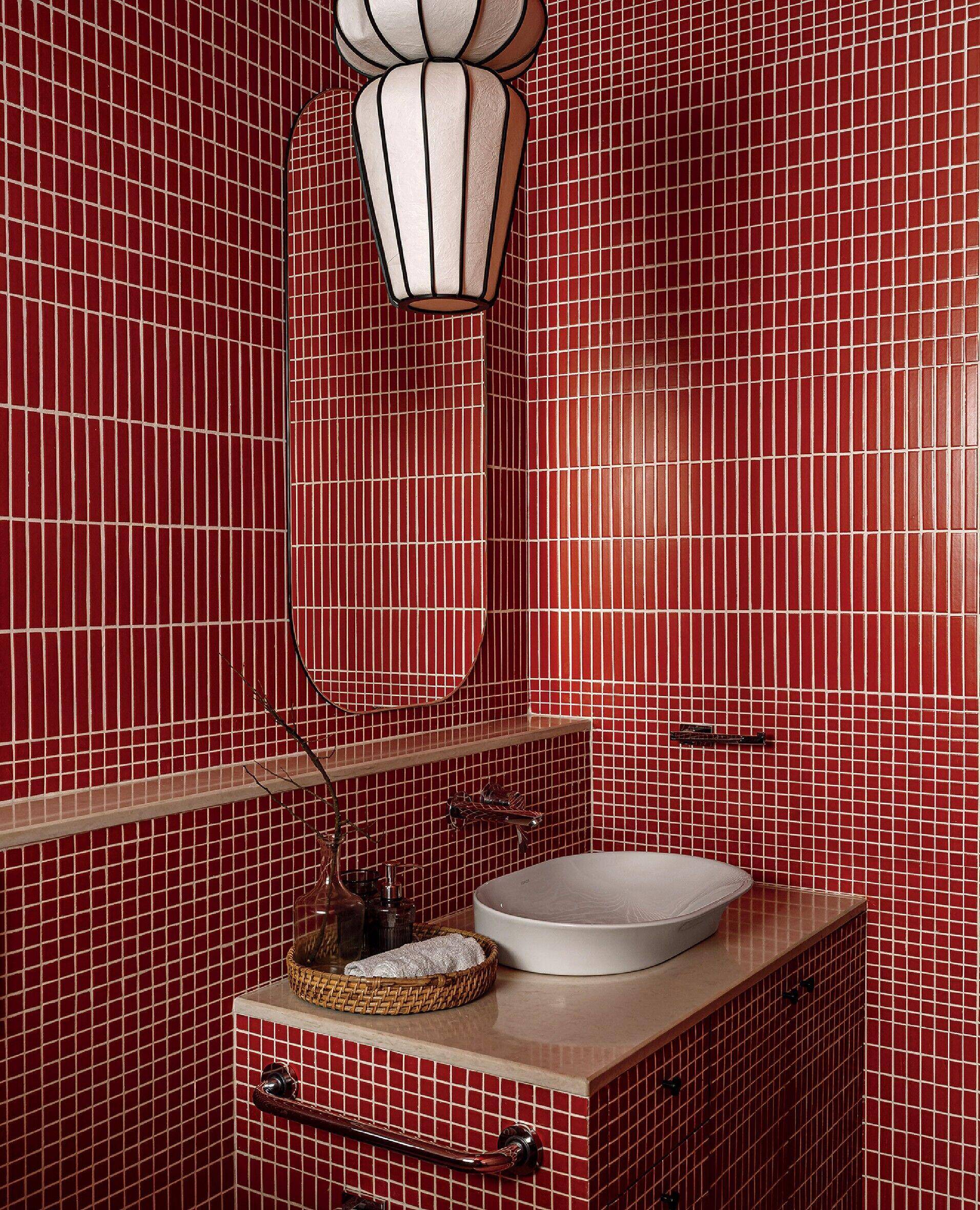 Small bathroom with red mosaic tile walls, a white vessel sink on a beige countertop, a woven tray with toiletries, and an oval mirror below a white pendant light.