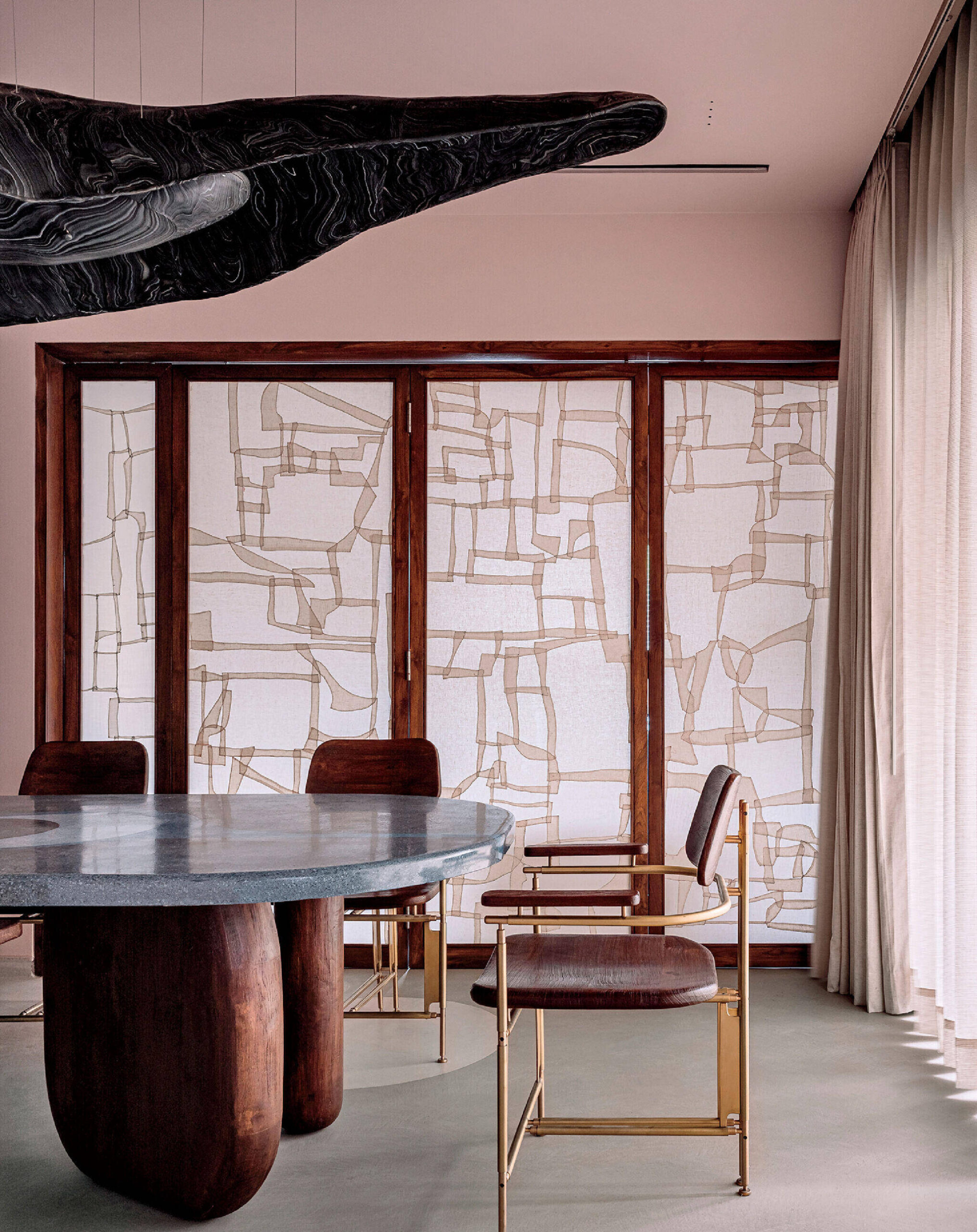 Modern dining room with a large oval table, four chairs with metal frames, abstract wall art, pink walls, and floor-to-ceiling curtains.