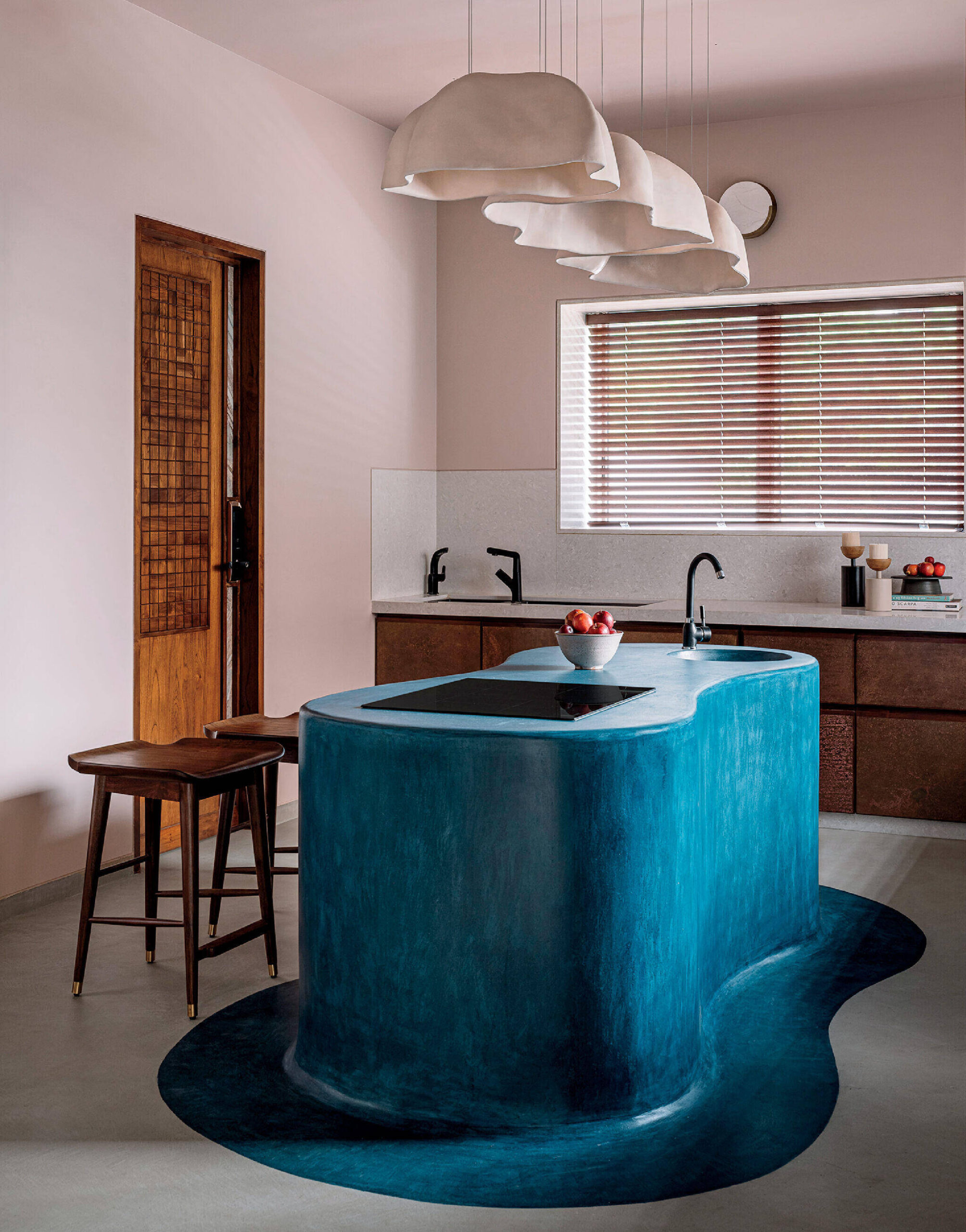 Modern kitchen with a sculpted blue island, wooden stools, and pendant lights. Brown cabinets, a window with blinds, and minimal decor complete the space.