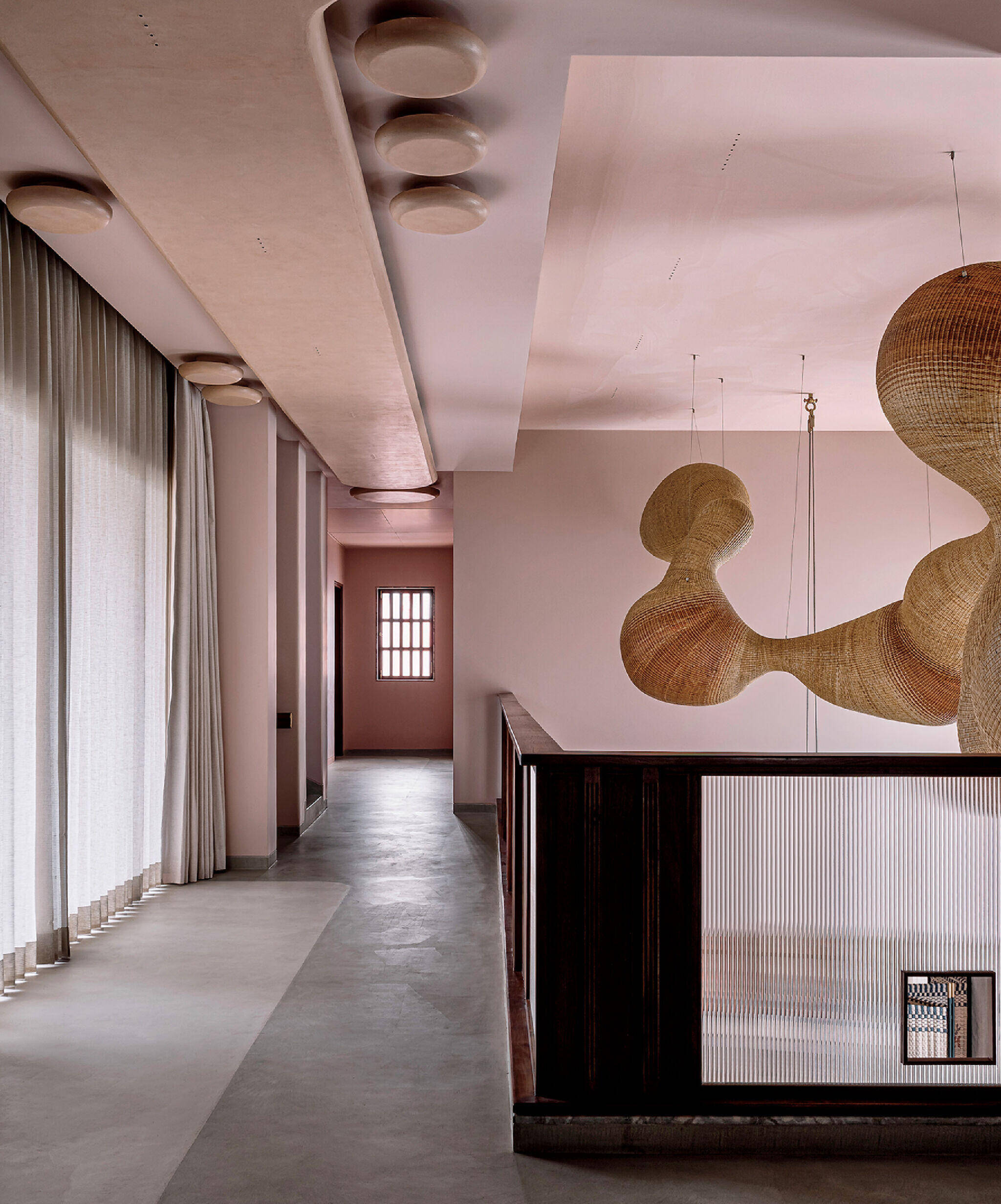 A modern hallway with light pink walls, large windows with sheer curtains, gray concrete floors, and sculptural woven pendant lights hanging from the ceiling.