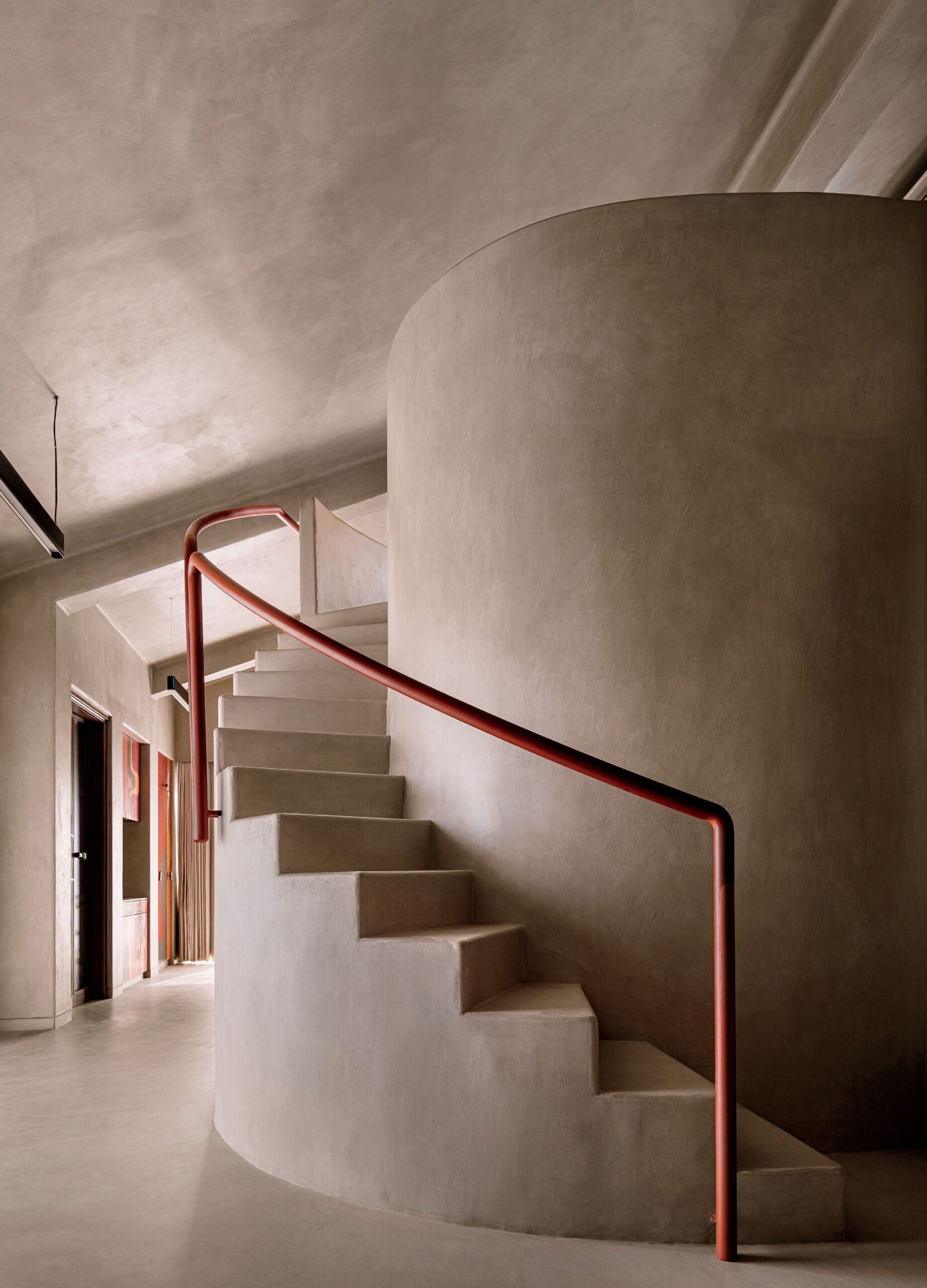 Curved concrete staircase with a smooth finish and a red handrail, set against beige walls in a modern interior space.