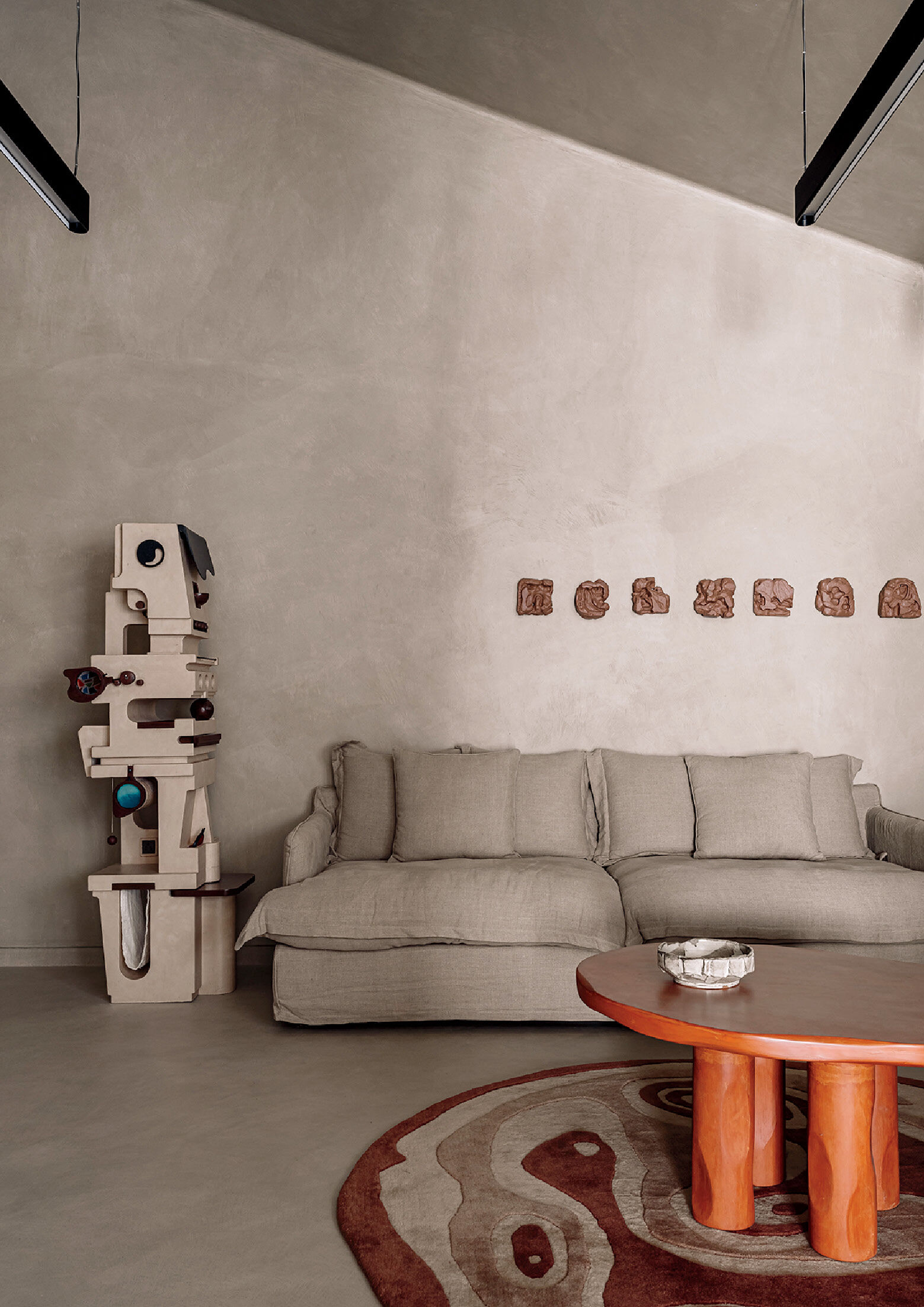 Minimalist living room with a beige sofa, sculptural decor piece, round wooden coffee table, abstract rug, and small wall art pieces on a textured beige wall.