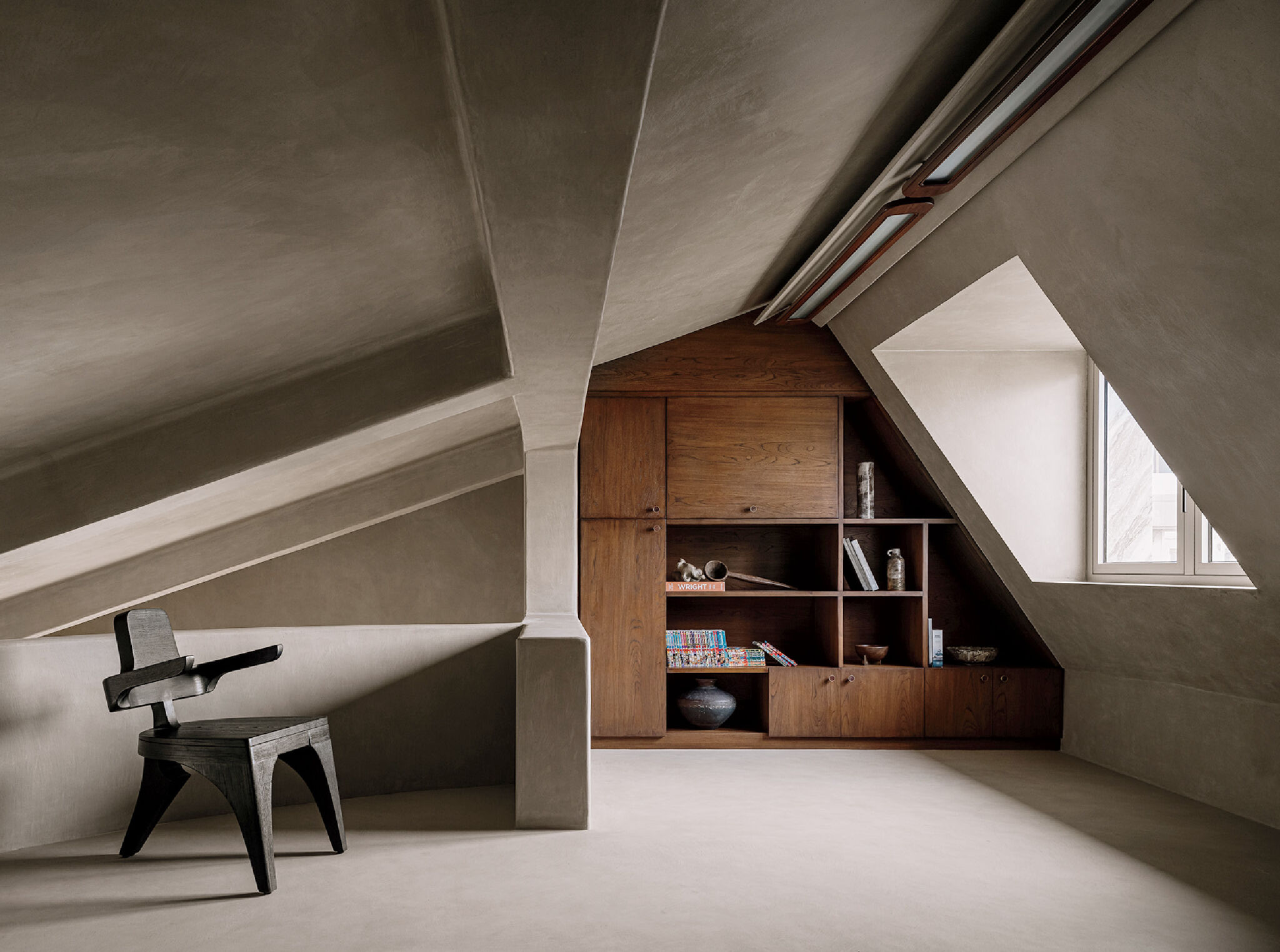Minimalist attic room with angled ceilings, a single modern black chair, built-in wooden shelves, books, and a window letting in natural light.