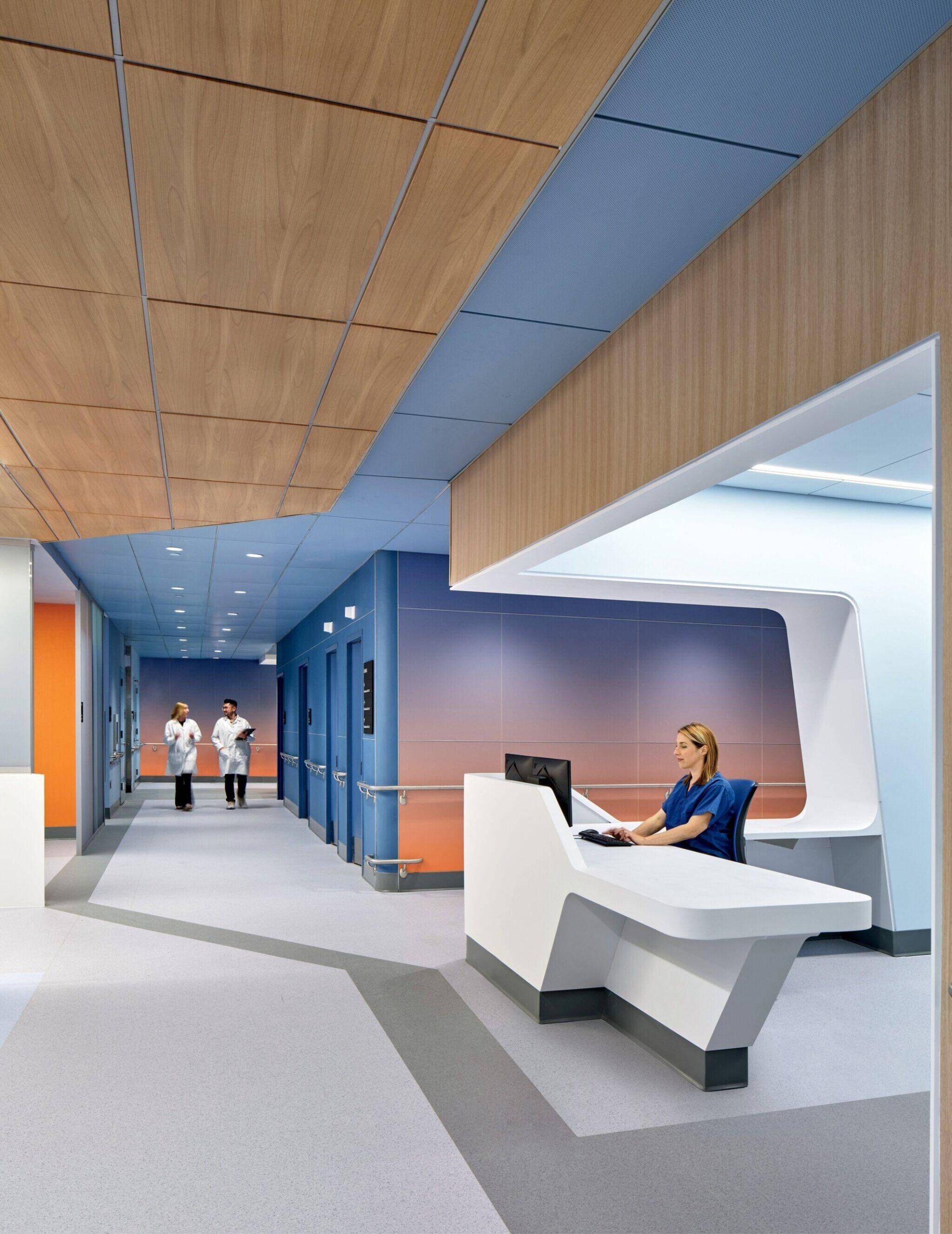 A nurse sits at a modern reception desk in a brightly lit hospital corridor as two medical staff walk down the hallway in the background.